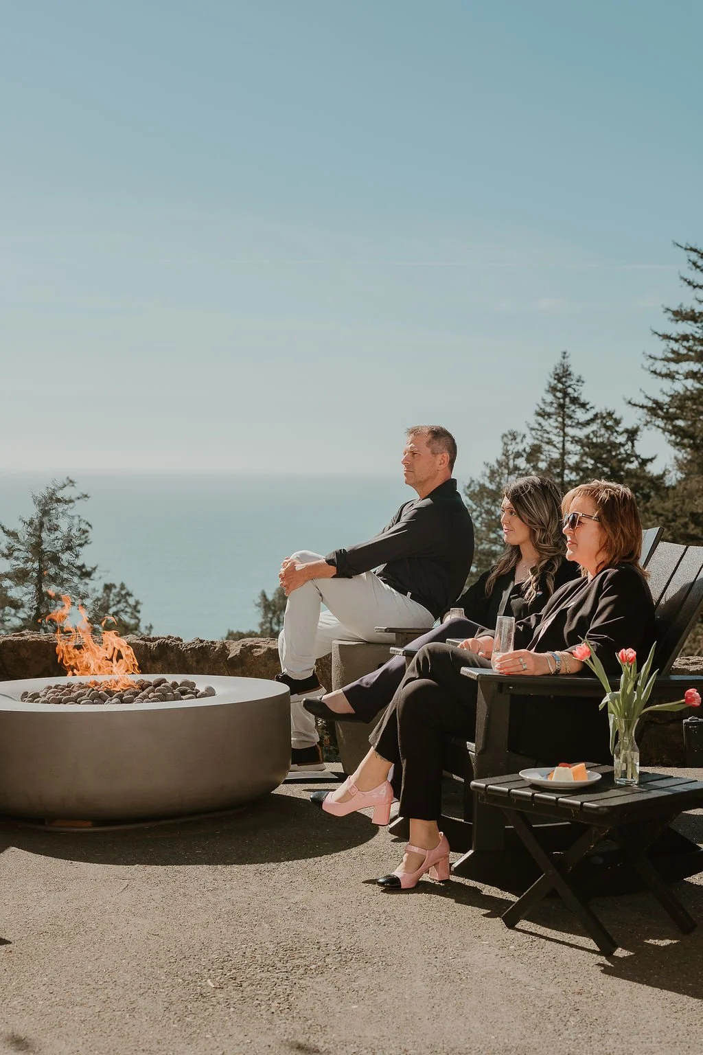 Three people sit outdoors on chairs near a fire pit with a view of trees and ocean in the background. One man and two women are seated, with the women holding glasses, and pastries on a small table nearby. The woman on the right wears sunglasses and pink heels, while the others are dressed casually.