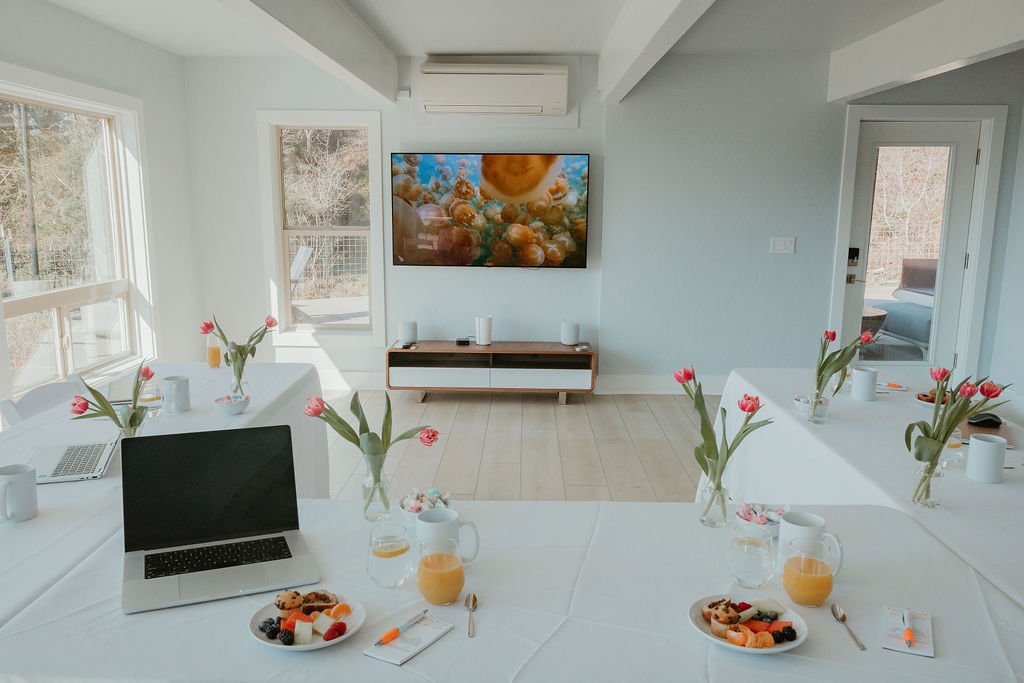 A bright, modern conference room set up for a meeting with white tablecloths, vases of pink tulips, laptops, plates of pastries, pitchers of orange juice, and cups. There is a television on the wall displaying a colorful image, and outside windows show a wooded area.