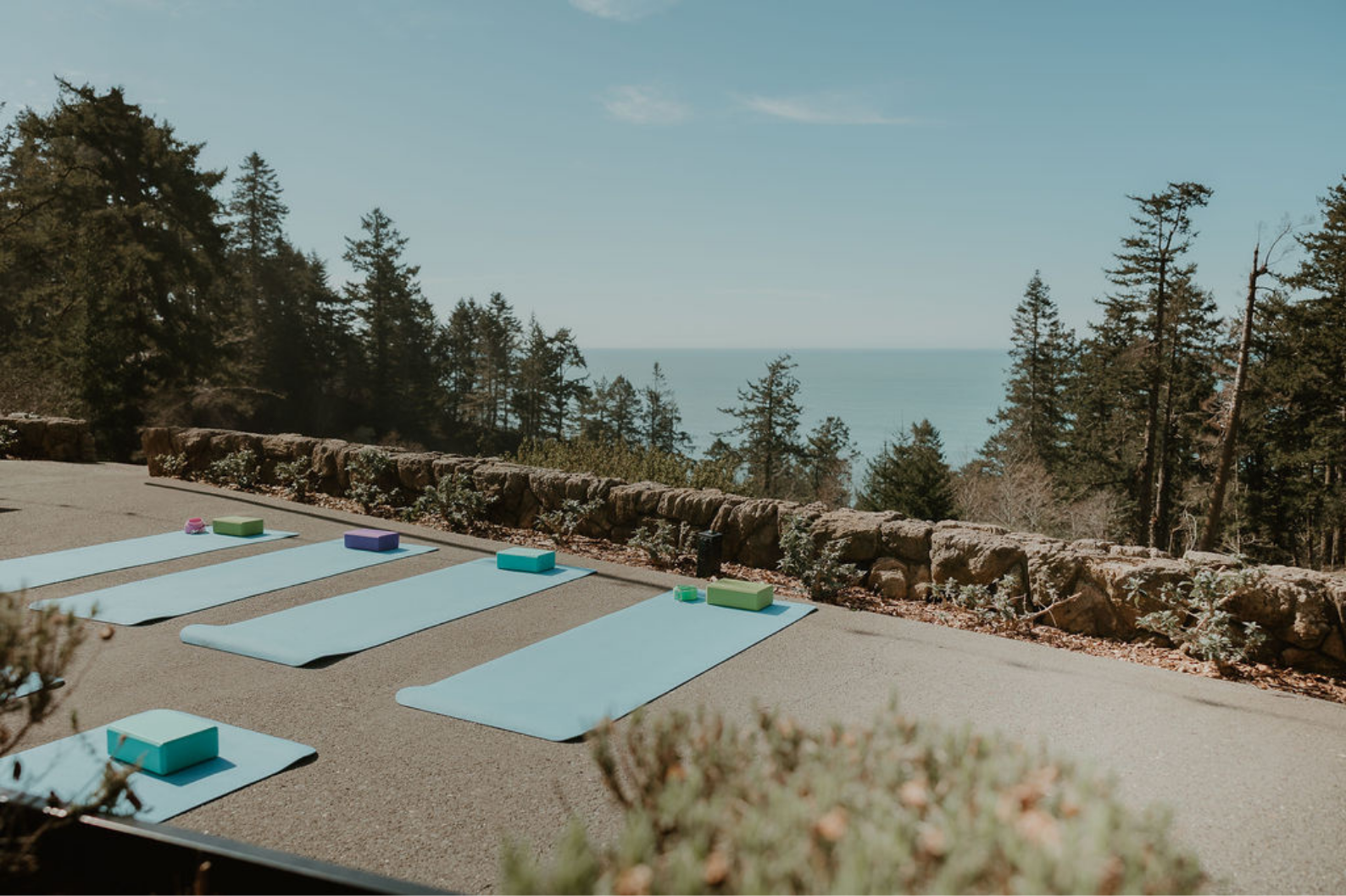 Outdoor yoga setup with light blue mats and colorful blocks on a paved surface, overlooking a forest and ocean in the distance under a clear sky.