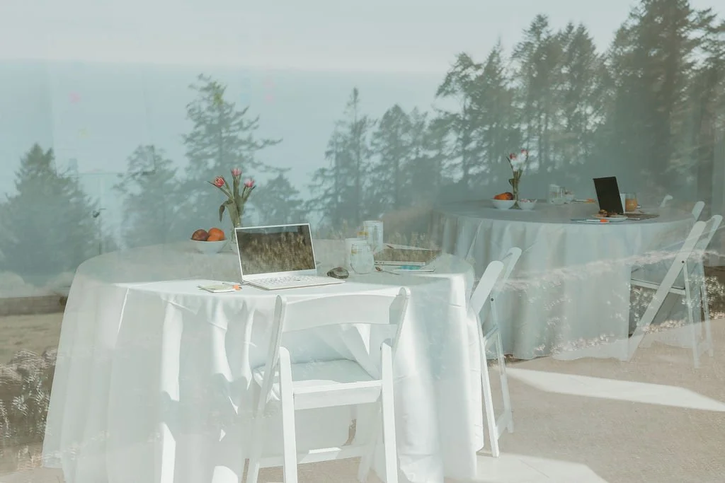 A reflection of a dining setup with round tables, white tablecloths, chairs, vases with pink flowers, laptops, and plates, seen through a glass window with a backdrop of trees and sky.