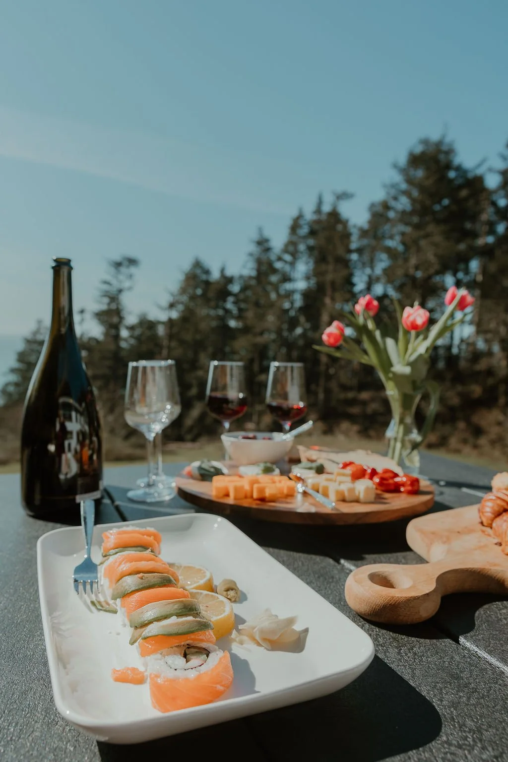 Sushi rolls, cheese platter, and wine glasses on outdoor table with a vase of pink tulips, set against a forested landscape under a clear sky.