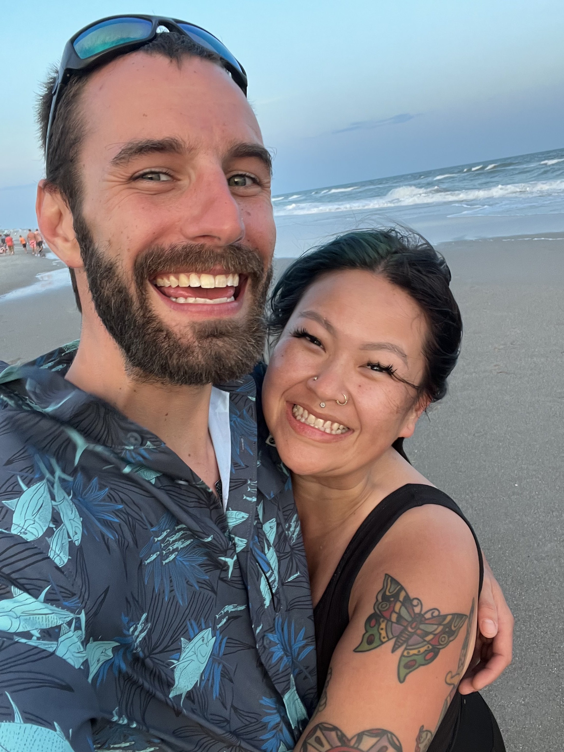A smiling man and woman taking a selfie on the beach with the ocean behind them.