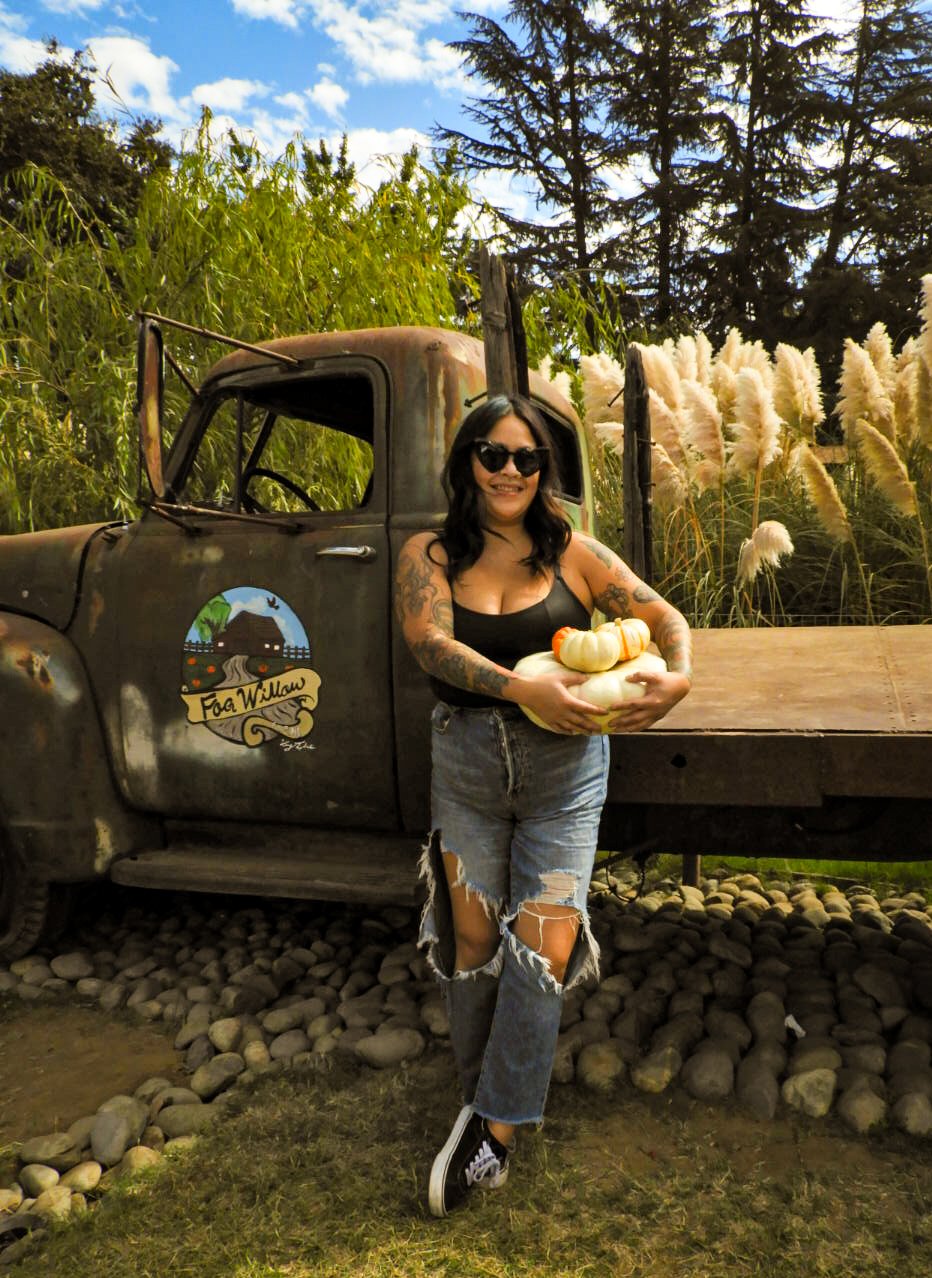 A woman with dark hair, sunglasses, and tattoos on her arms holding a pumpkin, standing next to an old, rusty truck with a farm logo. She is wearing a black tank top and ripped jeans, with a background of green trees and tall, white grass.