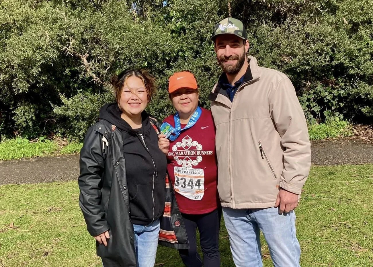 Three people standing together outdoors in front of greenery, smiling. The person in the middle is wearing a marathon shirt, has a race bib, and is holding a finisher medal. The other two are dressed casually, one with a jacket and the other with a c