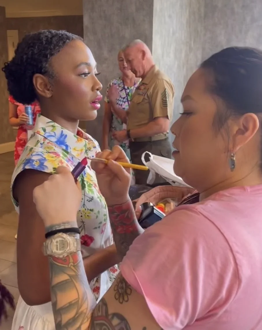 A woman with tattoos is applying lipstick to a young girl during a public event, with Marine staff and other people in the background.