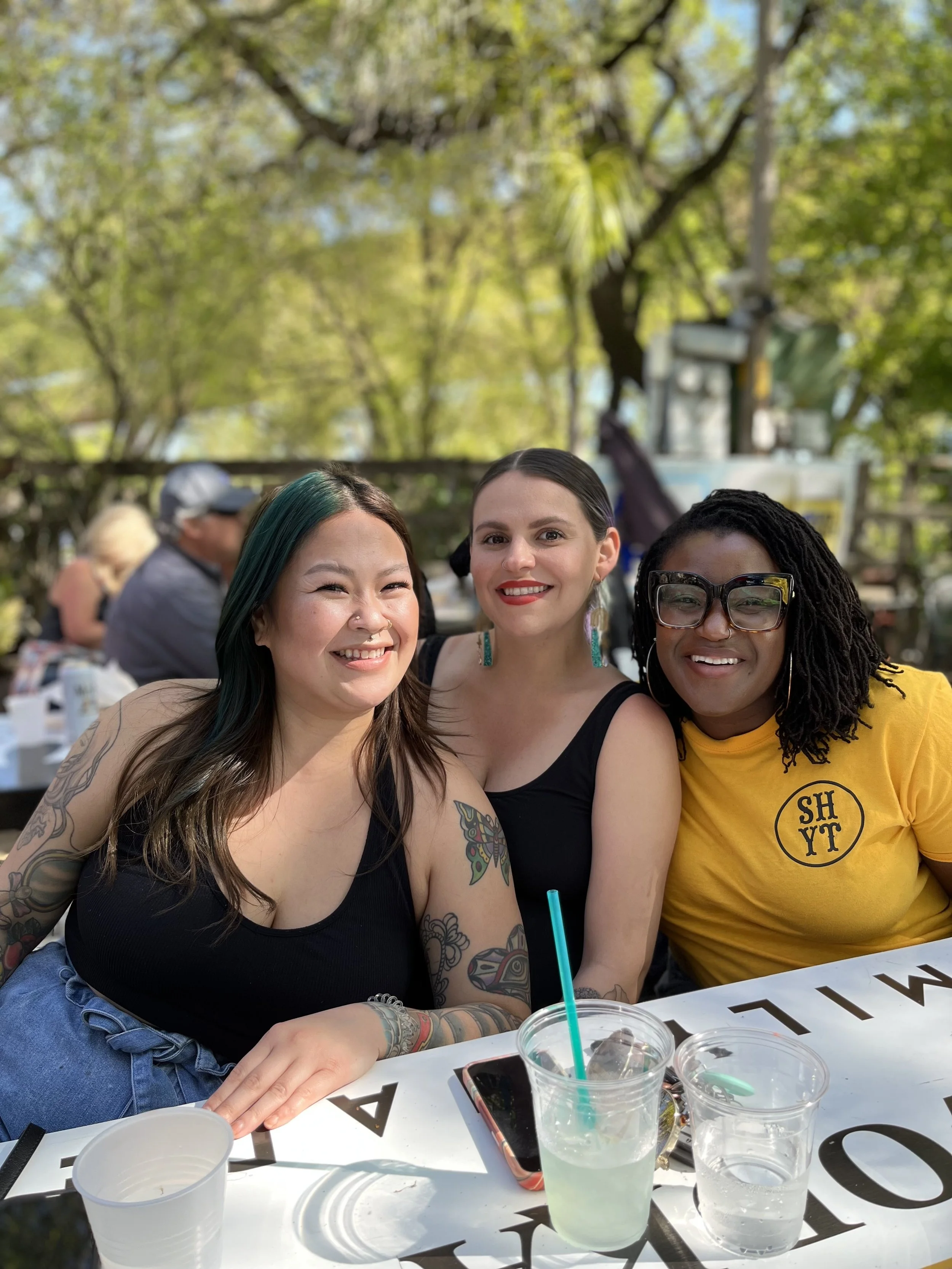 Three women smiling at an outdoor gathering, sitting at a table with drinks and tableware, with green trees in the background on a sunny day.