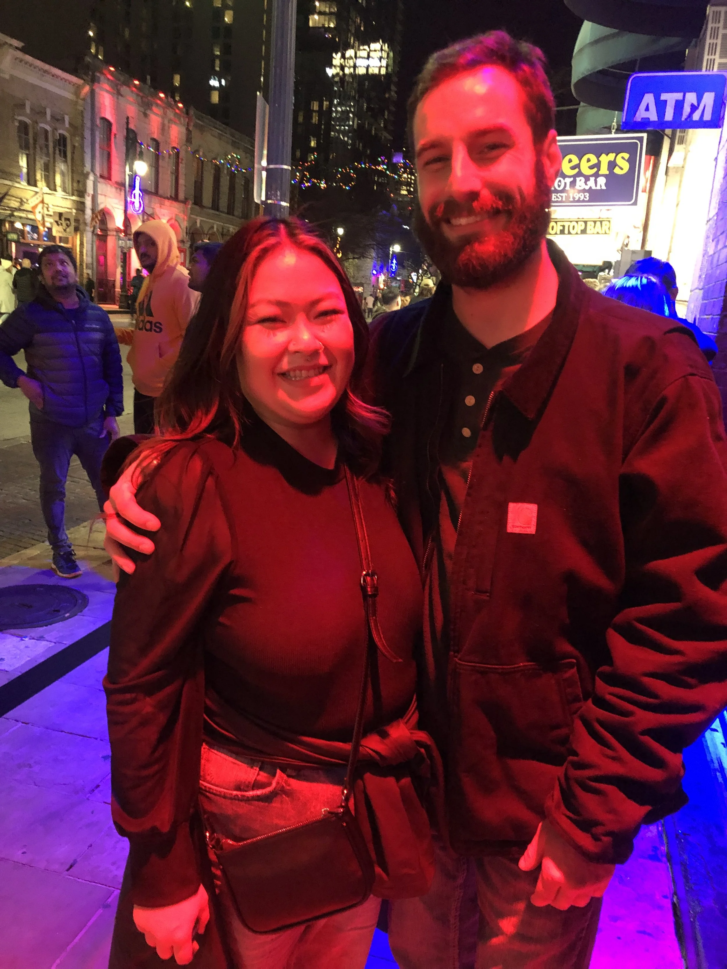 Two smiling people, a woman and a man, pose closely together on a city street at night. The background shows a bar and colorful city lights.