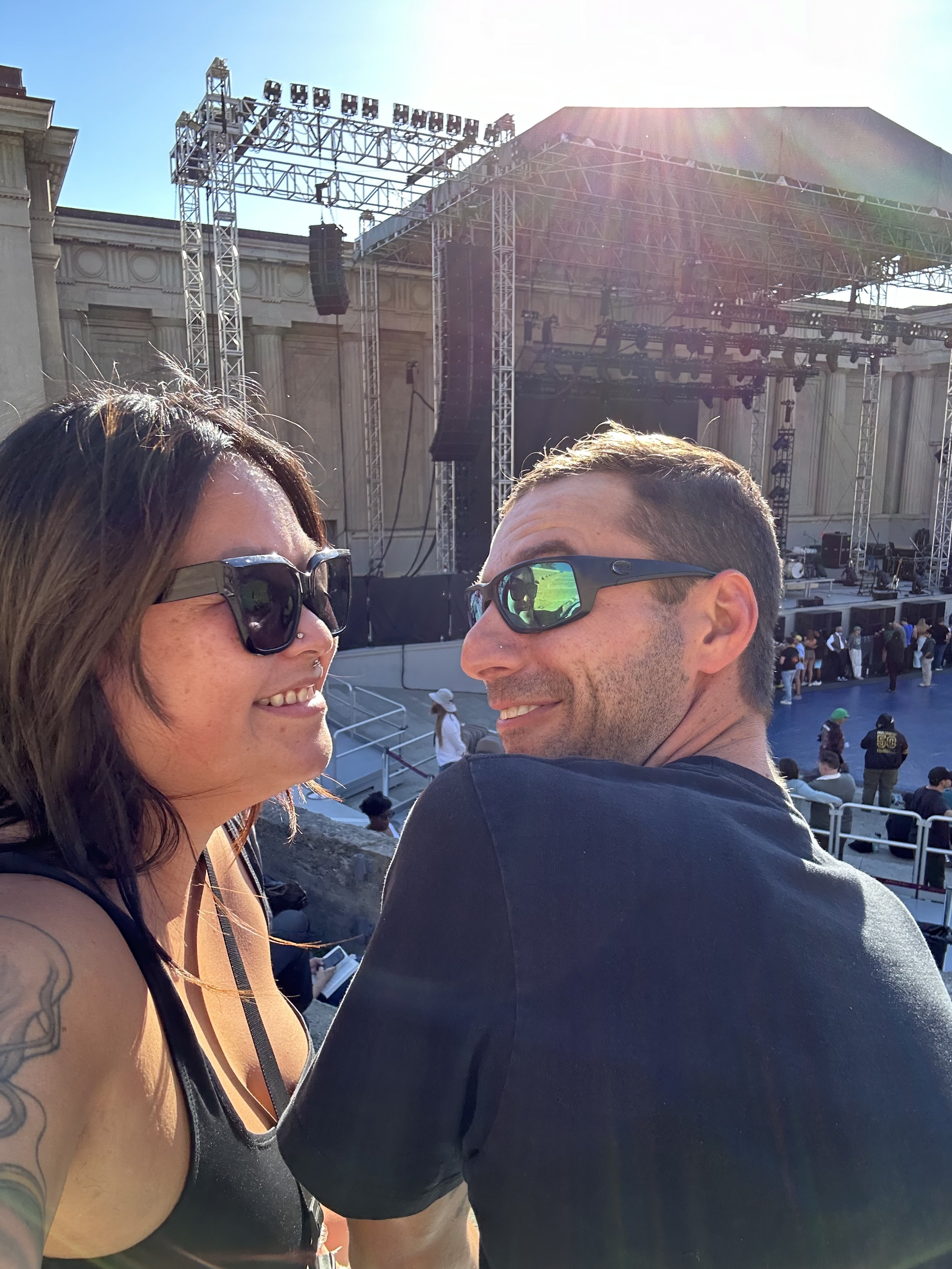 A woman and a man are close together, smiling, at an outdoor concert or event with a stage in the background and bright sunlight.