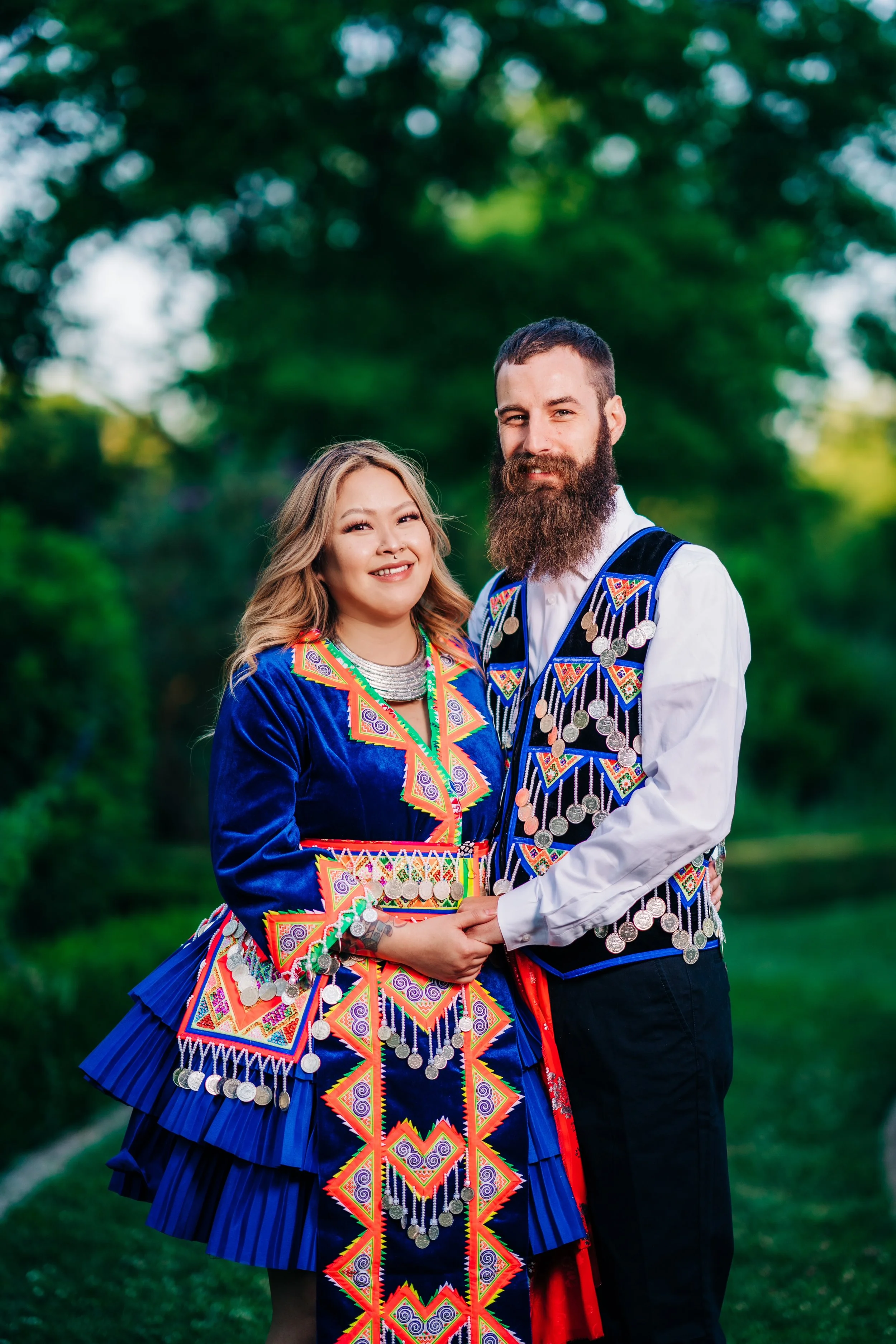 A man and woman dressed in traditional, colorful, beaded clothing with silver coins, standing outdoors with green trees in the background, smiling and holding hands.