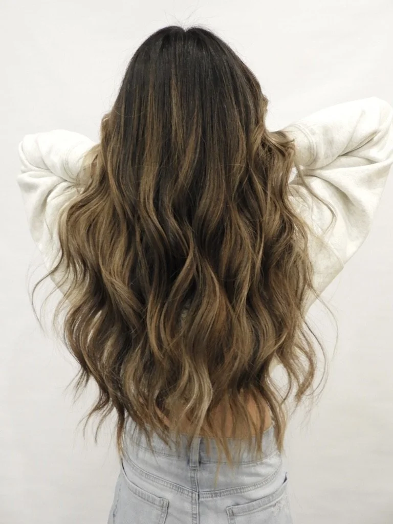 Back view of a woman with long, wavy, brown and blonde highlighted hair, wearing a white top and light-colored jeans, standing against a plain white wall.