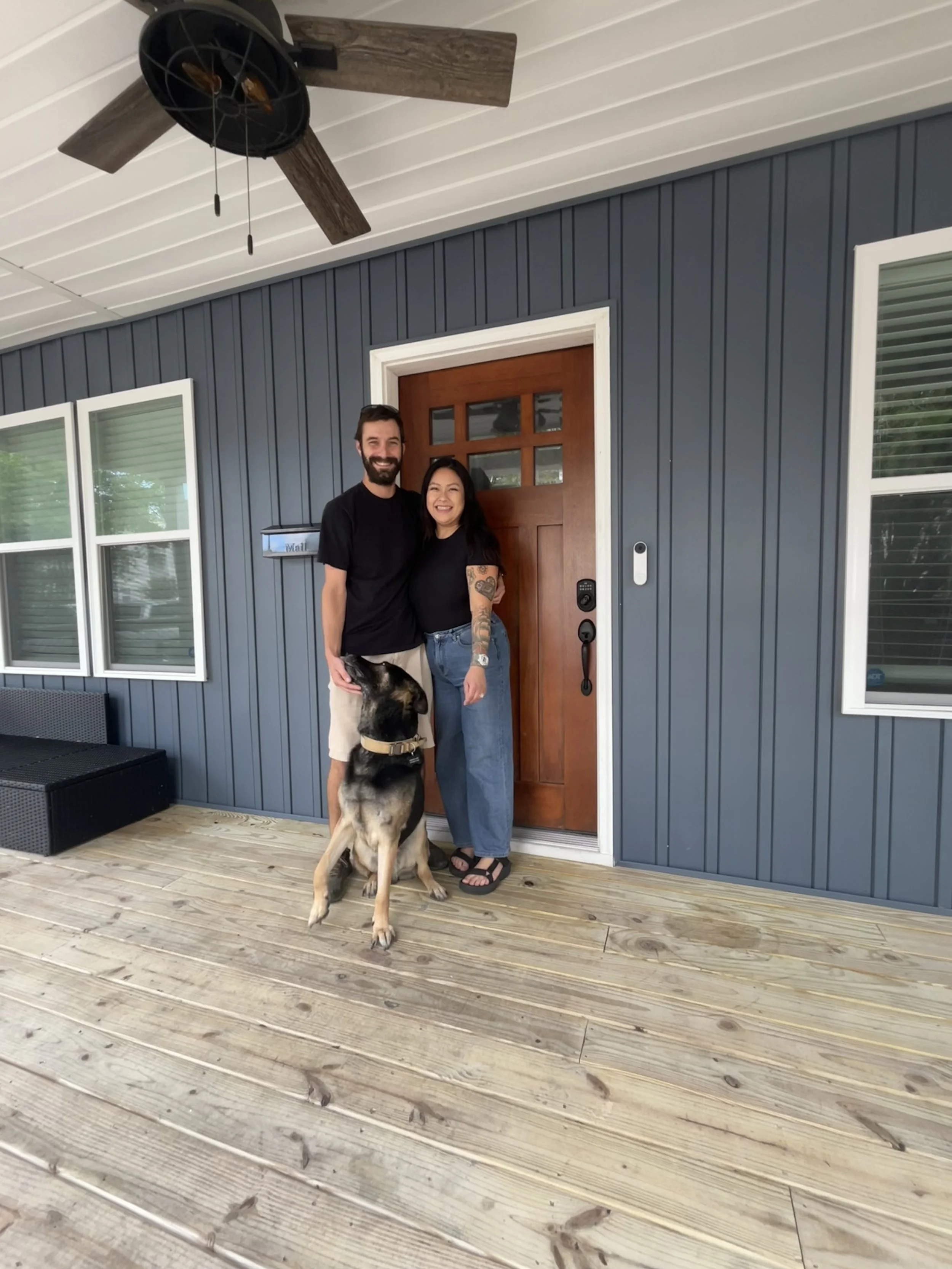 A smiling couple standing on a wooden porch in front of a blue house with a wooden door, with a large German Shepherd dog between them. The woman has long dark hair and tattoos on her arm, wearing a black shirt and blue jeans, and the man has short d
