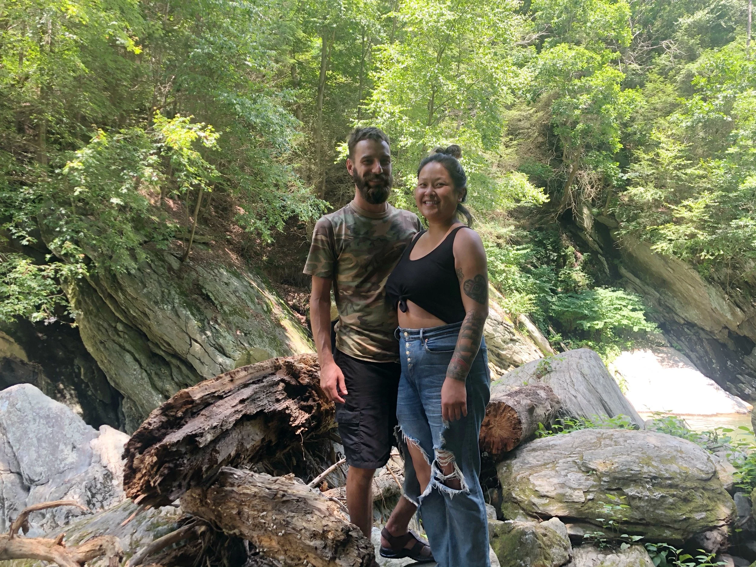 Two people, a man and a woman, standing outdoors on a rocky riverbank surrounded by green trees.