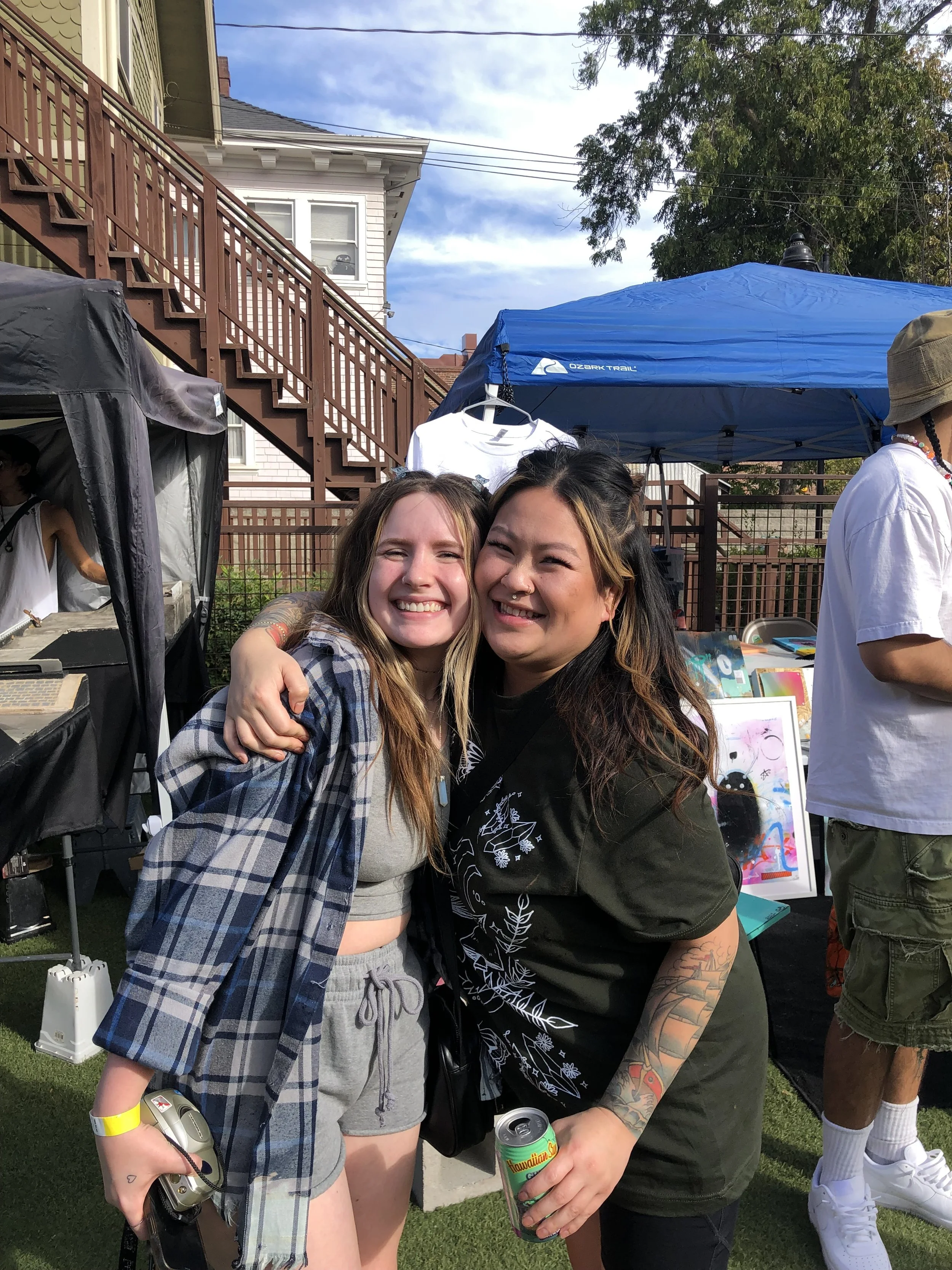 Two women smiling and embracing at an outdoor event with booths and a blue canopy in the background.
