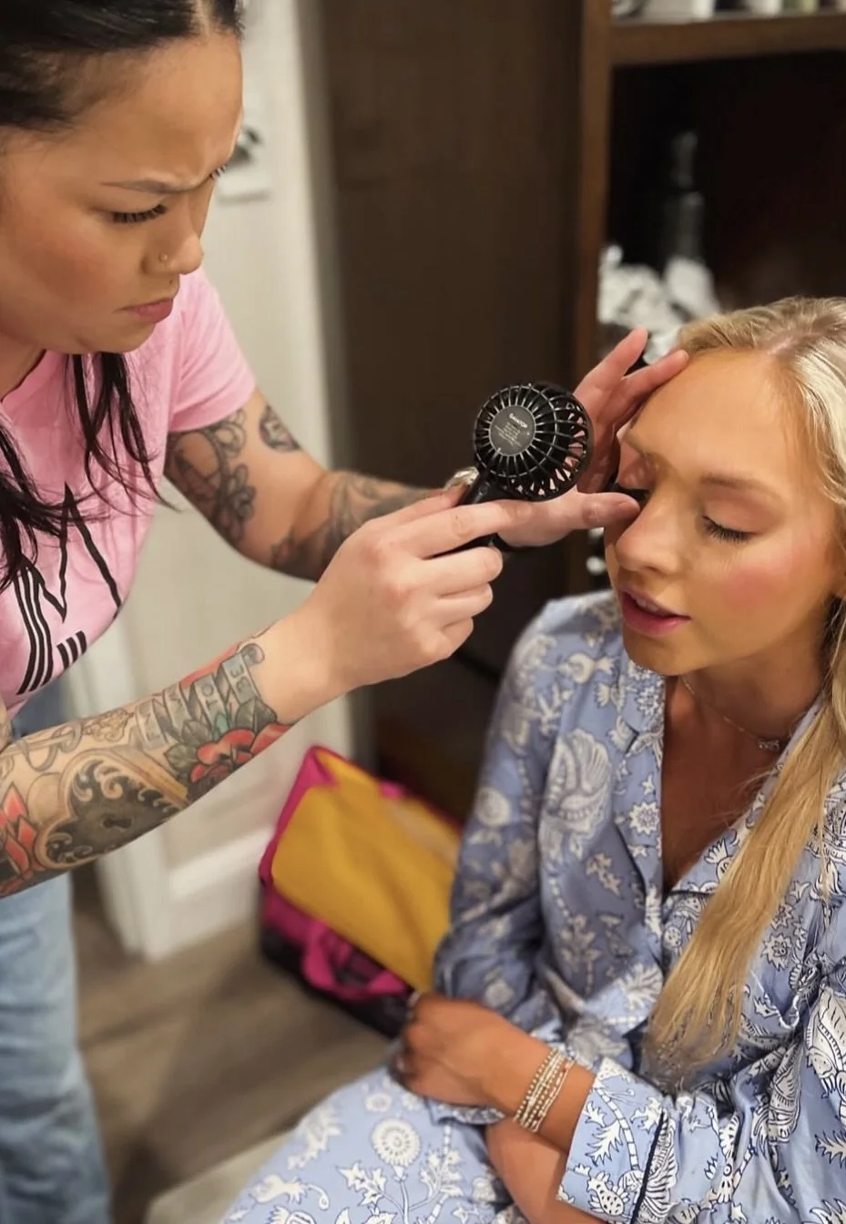 A woman with tattoos using a handheld fan on a woman's forehead.