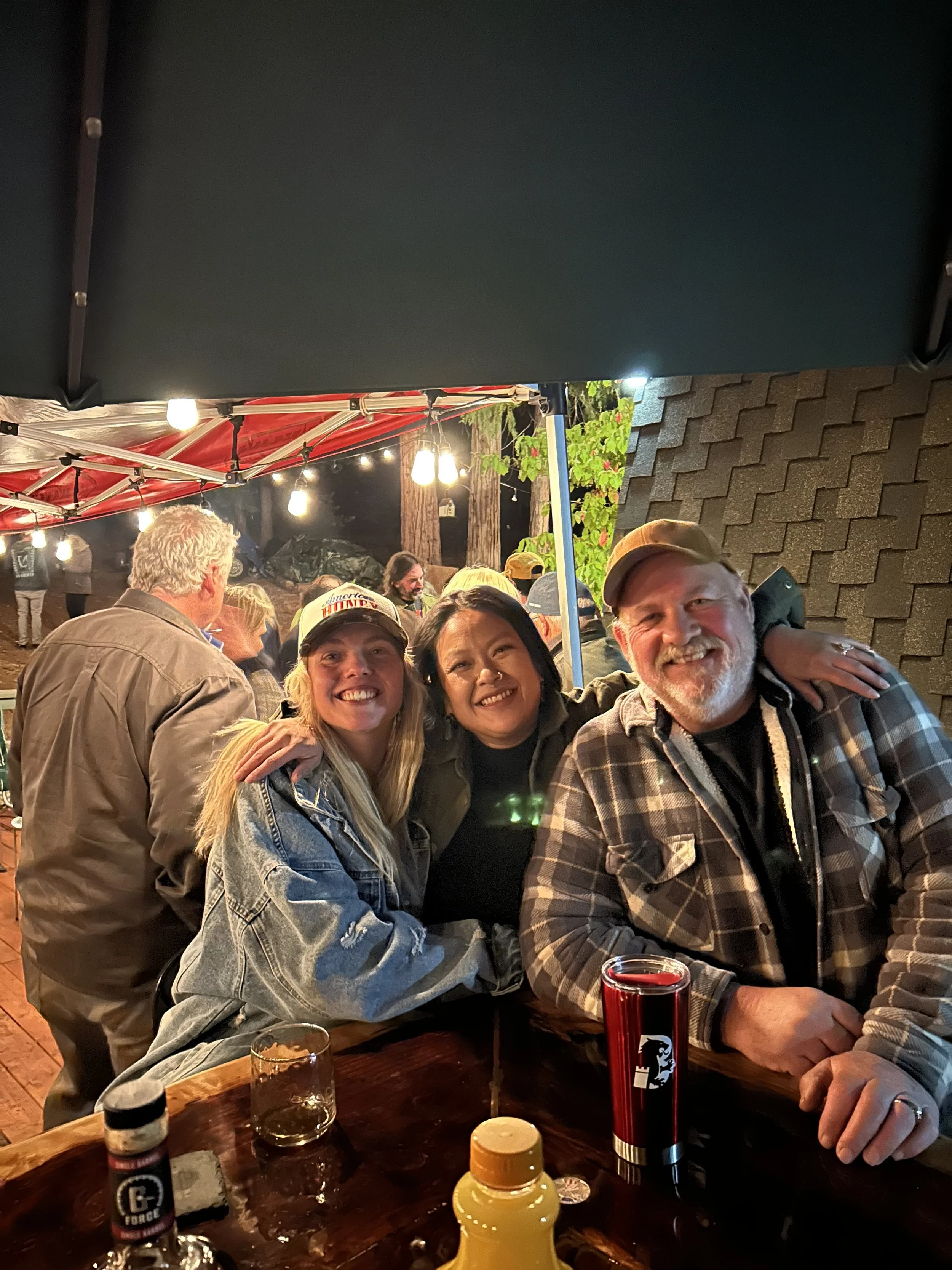 A group of people gathered at an outdoor night event, smiling and hugging, with a red canopy and hanging string lights overhead, and a table with drinks and condiments in front.