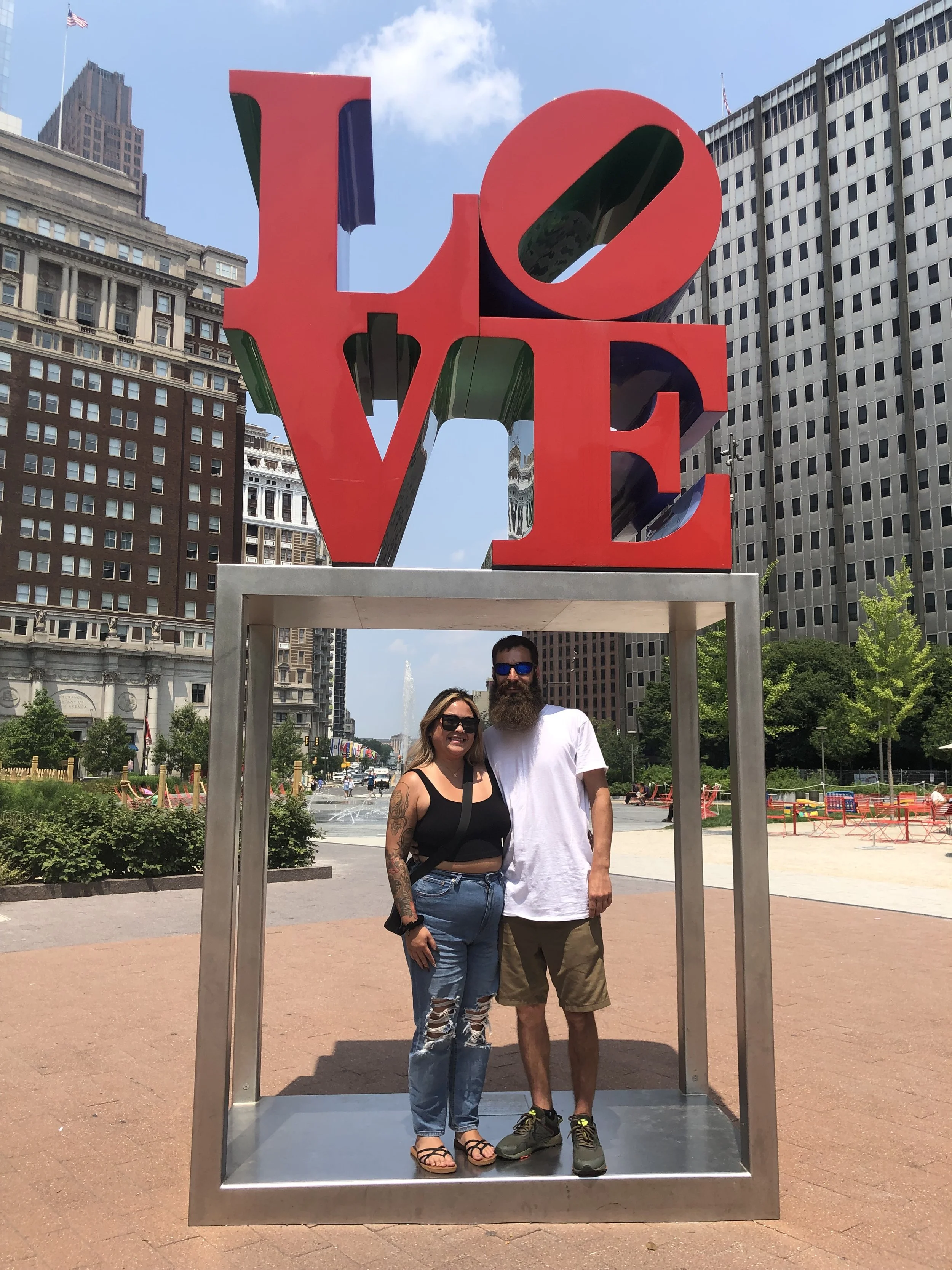 Two people standing inside a large multi-colored 'LOVE' sculpture with the cityscape and a fountain in the background.
