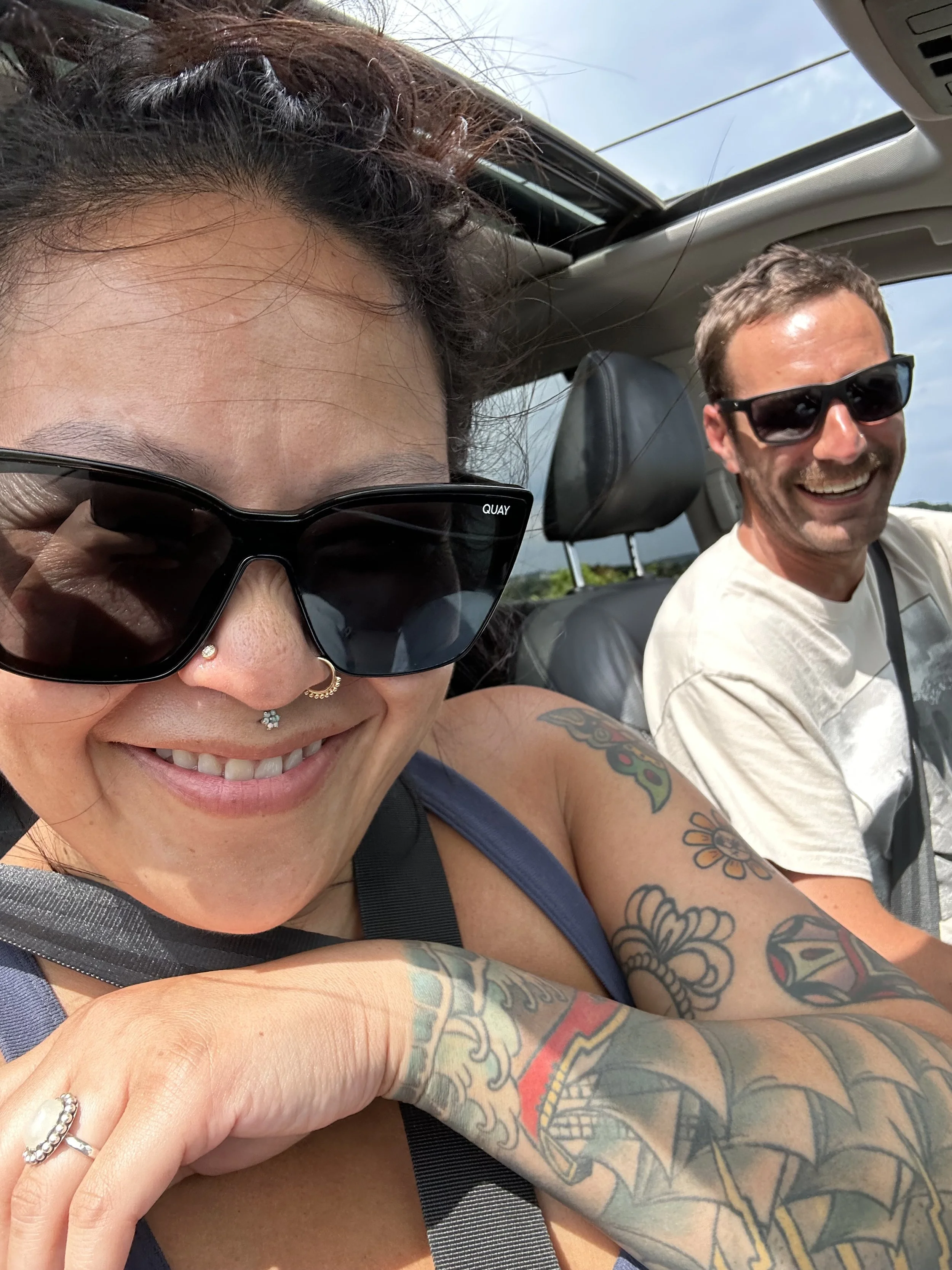 A woman and a man are taking a selfie inside a car, both wearing sunglasses and smiling. The woman has tattoos on her arms and face, and the man appears to be the driver.