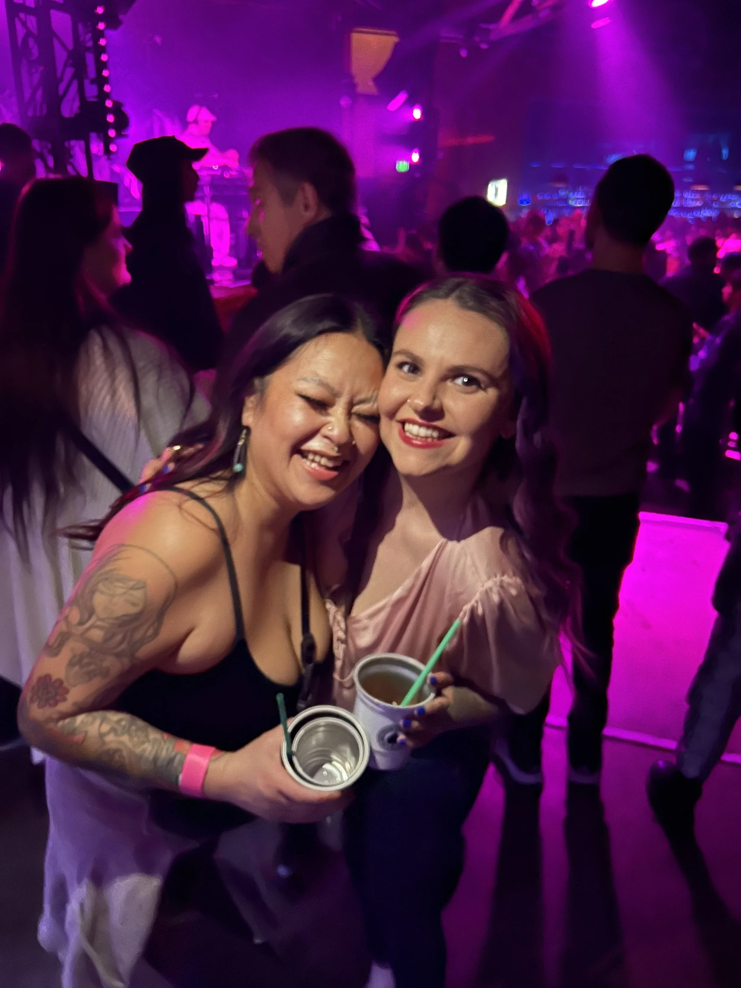 Two women smiling and holding drinks at a party or nightclub, with purple lighting and a DJ performing in the background.