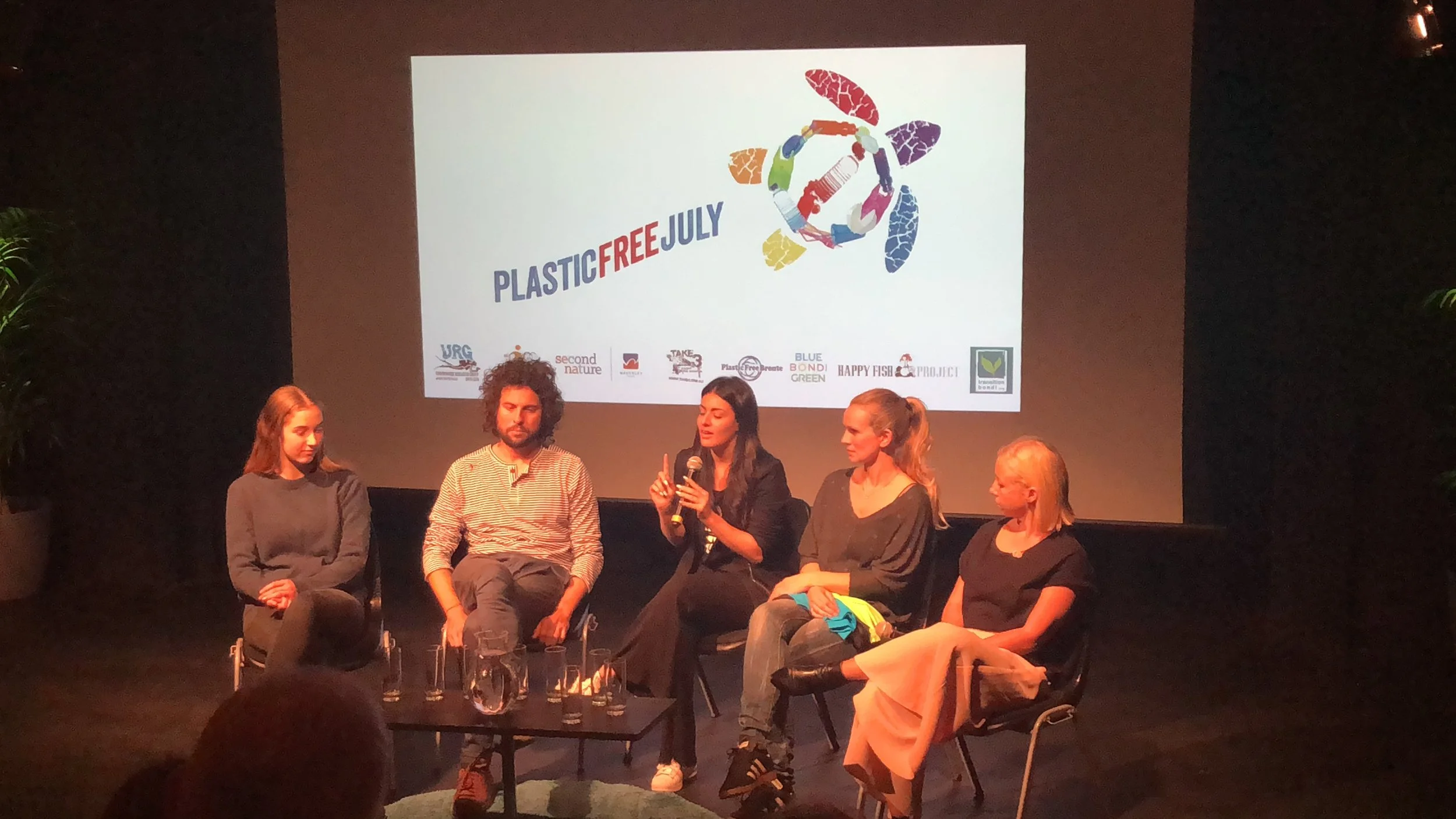 Five people sitting on a stage in front of a large screen with a logo and the words 'PLASTIC FREE JULY.' One woman is speaking into a microphone while the others listen.