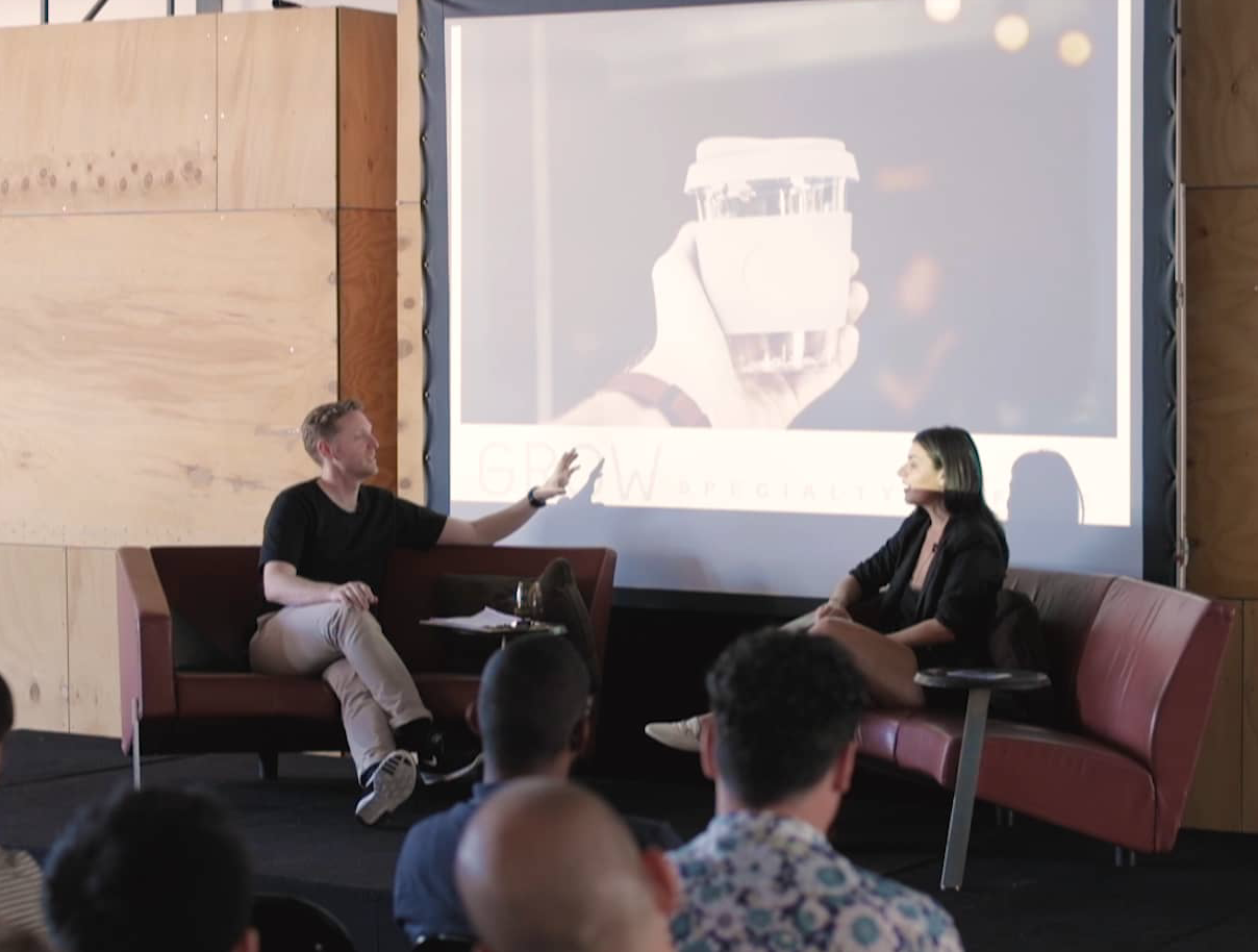 A man and a woman sitting on a stage in front of an audience, engaging in a discussion. The man is gesturing with his hand while speaking, and the woman is listening. Behind them is a large screen displaying an image of a hand holding a disposable coffee cup.