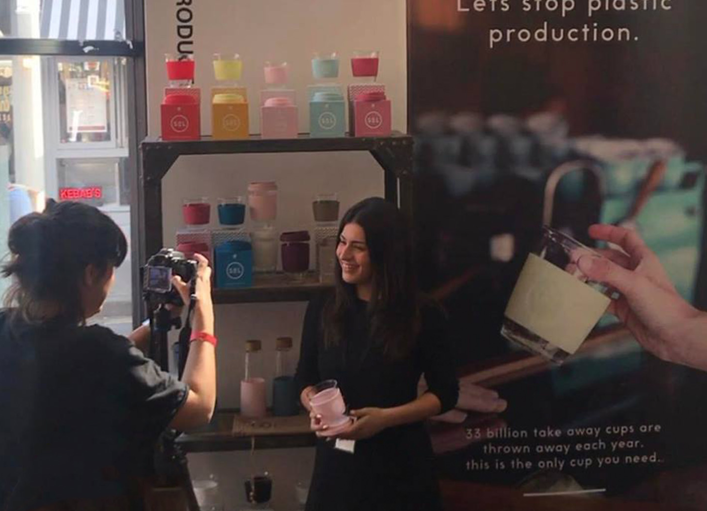 A woman with dark hair smiling and holding a pink cup while posing for a photo at a display of colorful cups inside a shop.