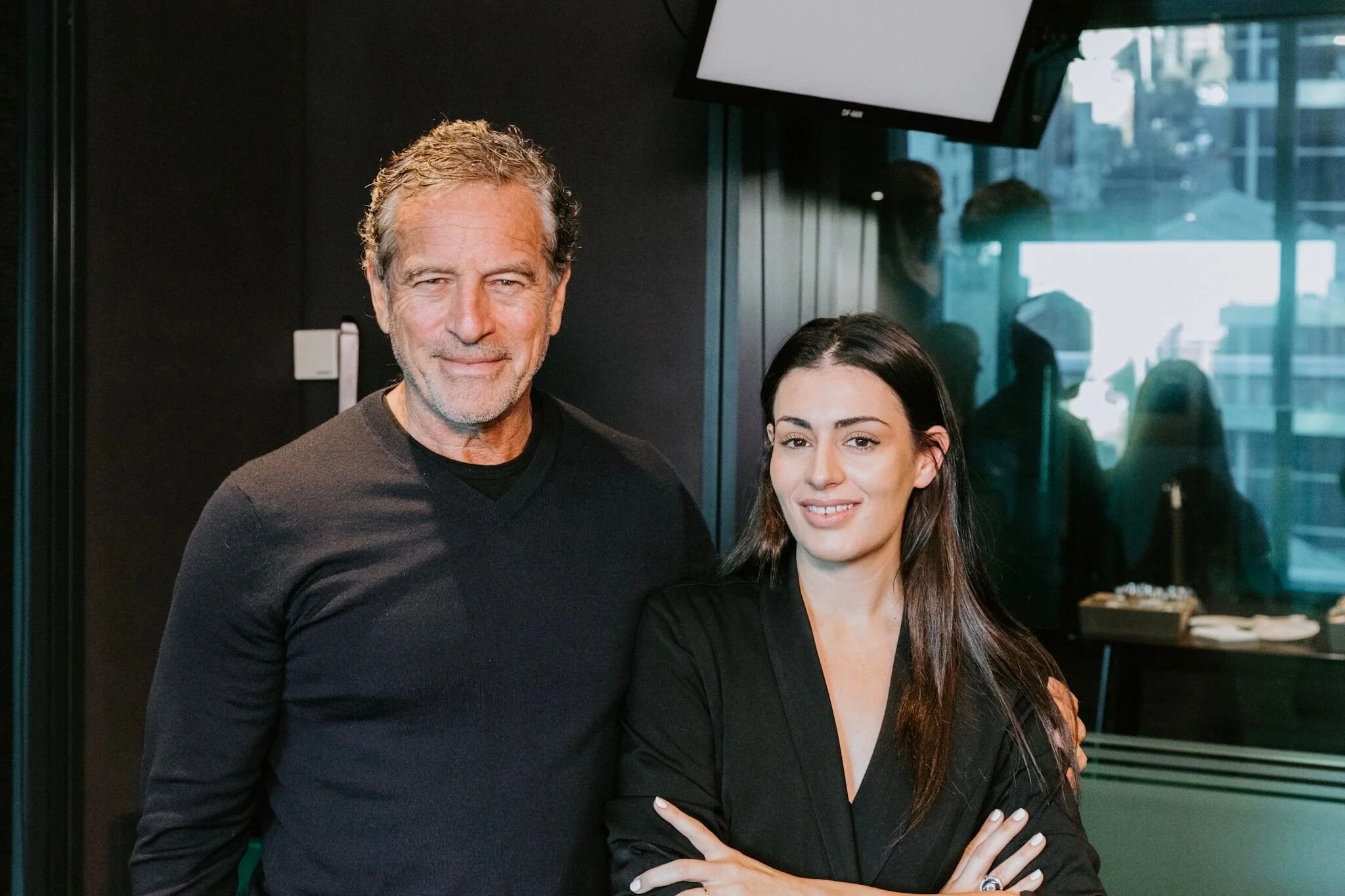 A man with gray hair and beard and a woman with long dark hair smiling and posing together indoors.