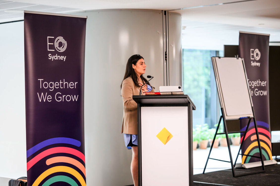 A woman giving a presentation at a conference or seminar, standing behind a podium with a microphone, in a room with banners displaying 'EO Sydney' and the slogan 'Together We Grow'.