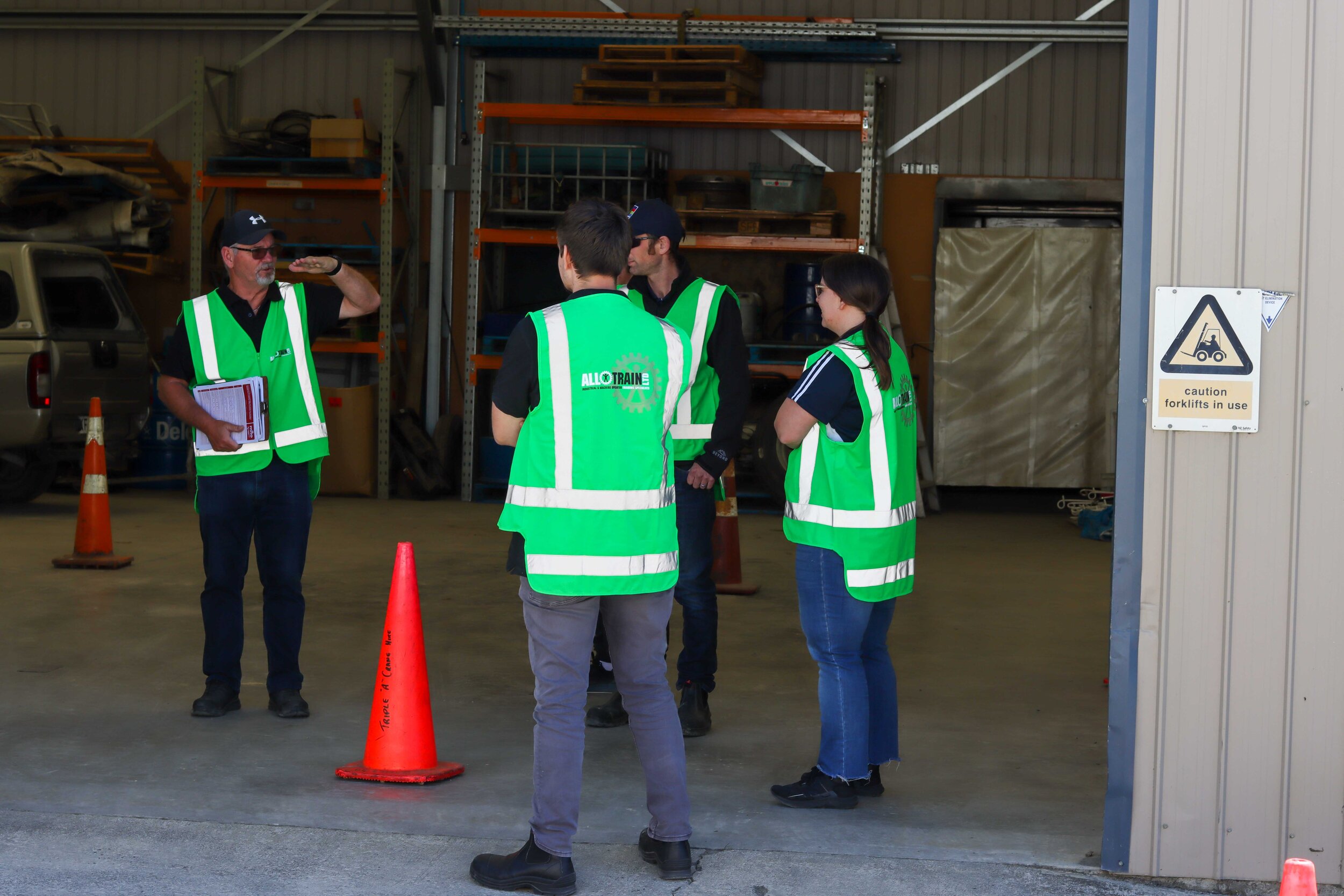 Group of four people wearing green safety vests inside a warehouse, discussing while a man salutes. Safety cones and a sign warning about forklift use are visible.