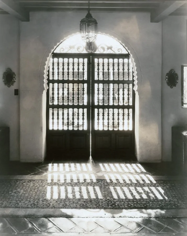  in a vintage photo, an ornate wooden grille casts shadows at an entrance to an interior patio. 