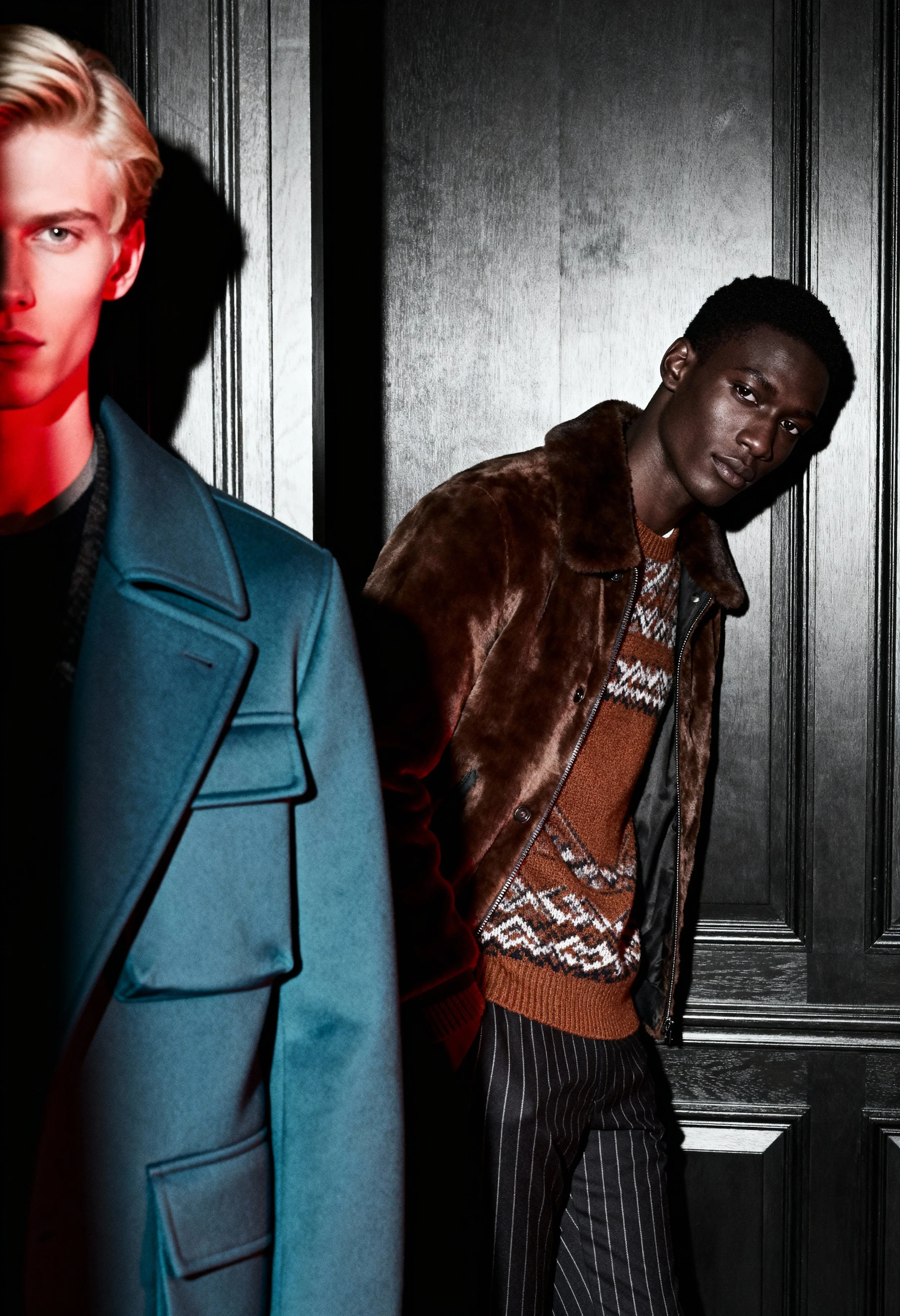 Two men standing against a dark wooden wall, one with blonde hair wearing a teal coat, and the other with dark skin and short curly hair wearing a brown fur coat over a patterned sweater and pinstriped pants.