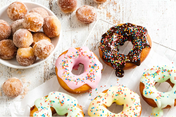 Assorted donuts with colorful sprinkles and glazed toppings, and sugar-coated donut holes in a bowl on a white surface.