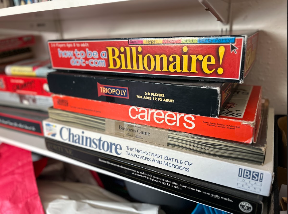 A shelf holding several board games, including 'How to be a Billionaire,' 'Triopoly,' 'Careers,' 'Chainstore,' and others, stacked one on top of the other.