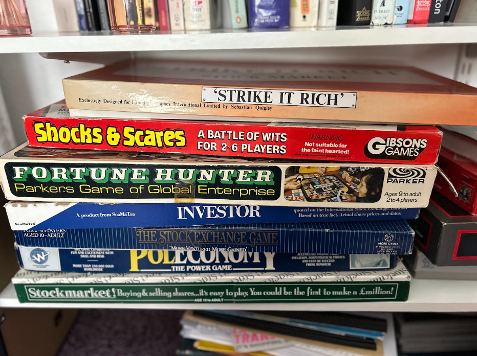 Stack of board game boxes on a shelf, including 'Strike It Rich,' 'Shocks & Scares,' 'Fortune Hunter,' 'Investor,' 'The Stock Exchange Game,' 'Pollonomy,' and 'Stockmarket!'