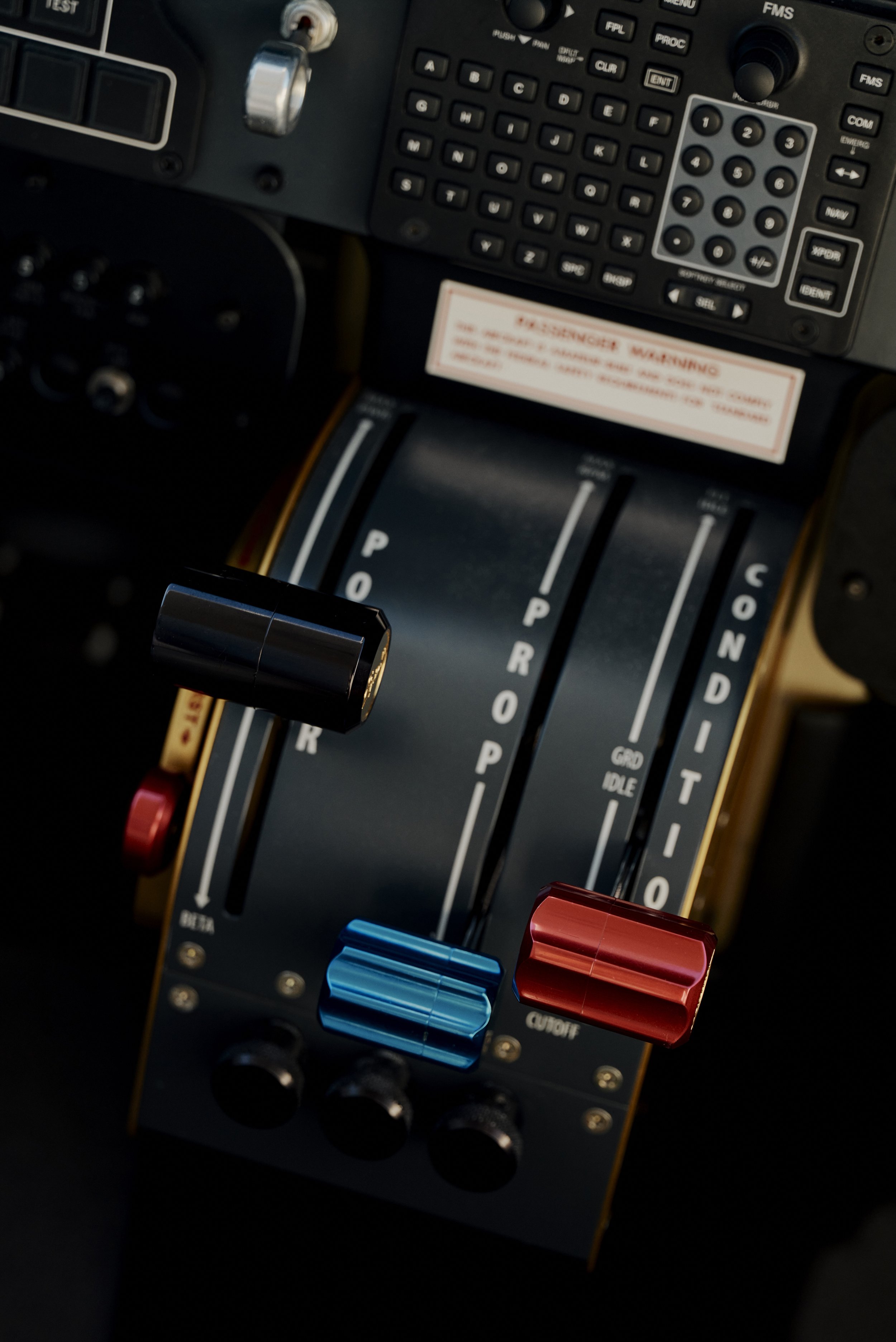 Nahaufnahme eines Flugzeugcockpit-Steuerhebels mit Schubreglern in verschiedenen Farben. Es sind verschiedene Bedienelemente und Aufschriften sichtbar, darunter "PROP" und "CONDITION". Ein Teil eines Bedienfelds mit Tasten ist ebenfalls zu sehen.