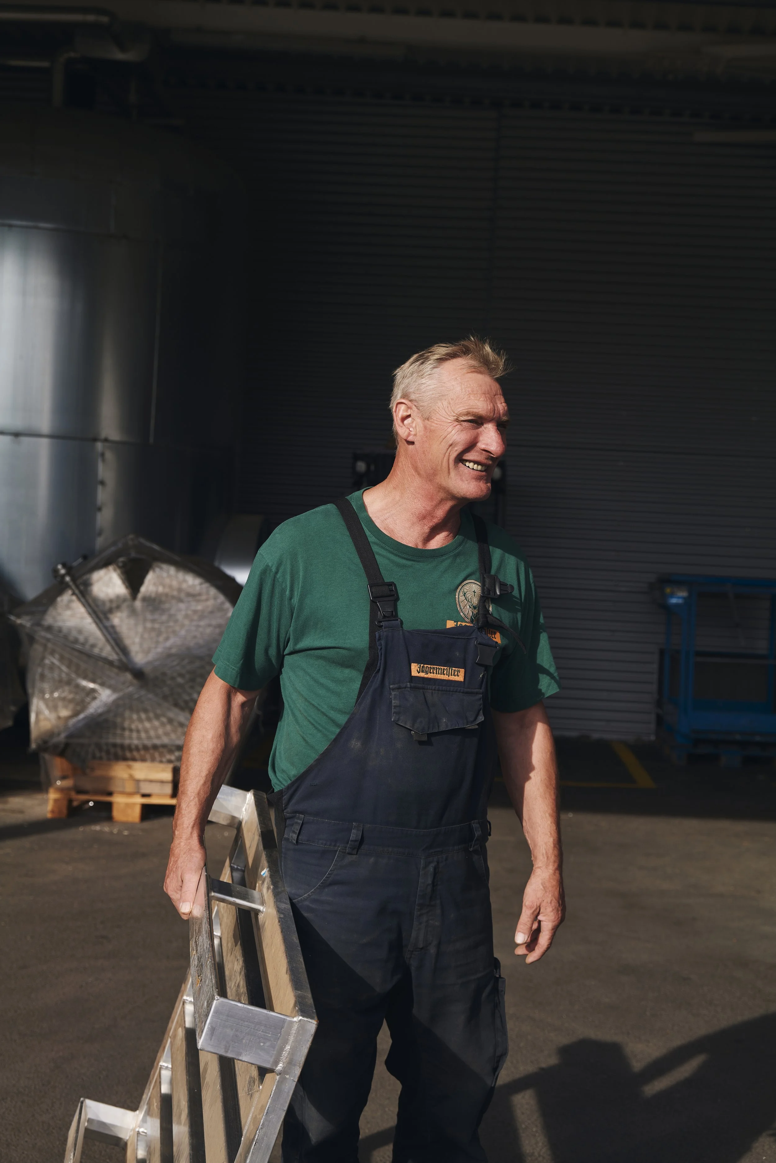 Mann in Arbeitskleidung mit einer Leiter im Freien, lächelnd neben einem Industriestandort.