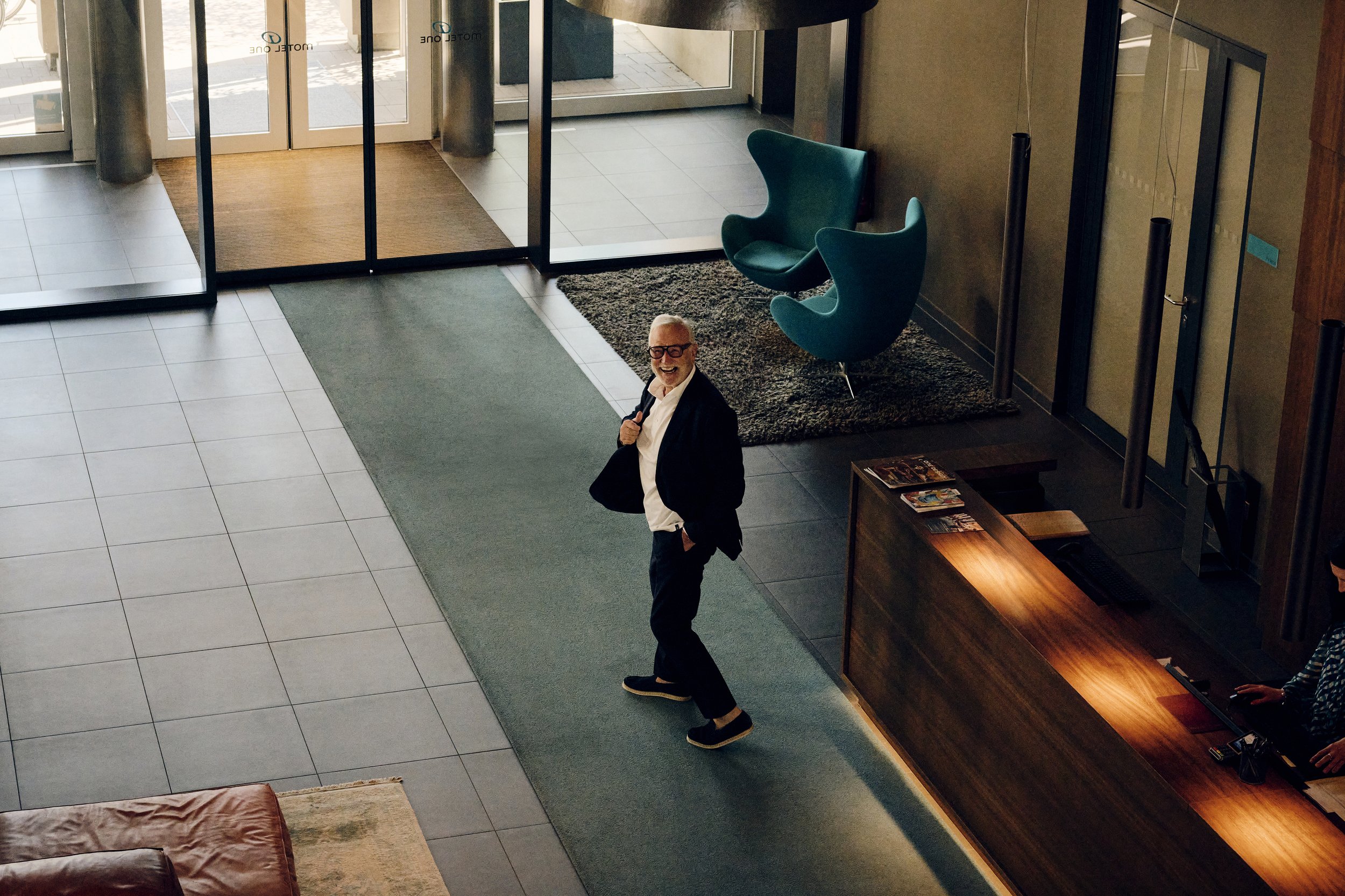 Hotel-Lobby mit Dieter Müller, der Richtung Rezeption geht. Moderne Einrichtung mit blauem Sessel auf Teppich. Empfangstresen im Vordergrund, Eingangstüren im Hintergrund.