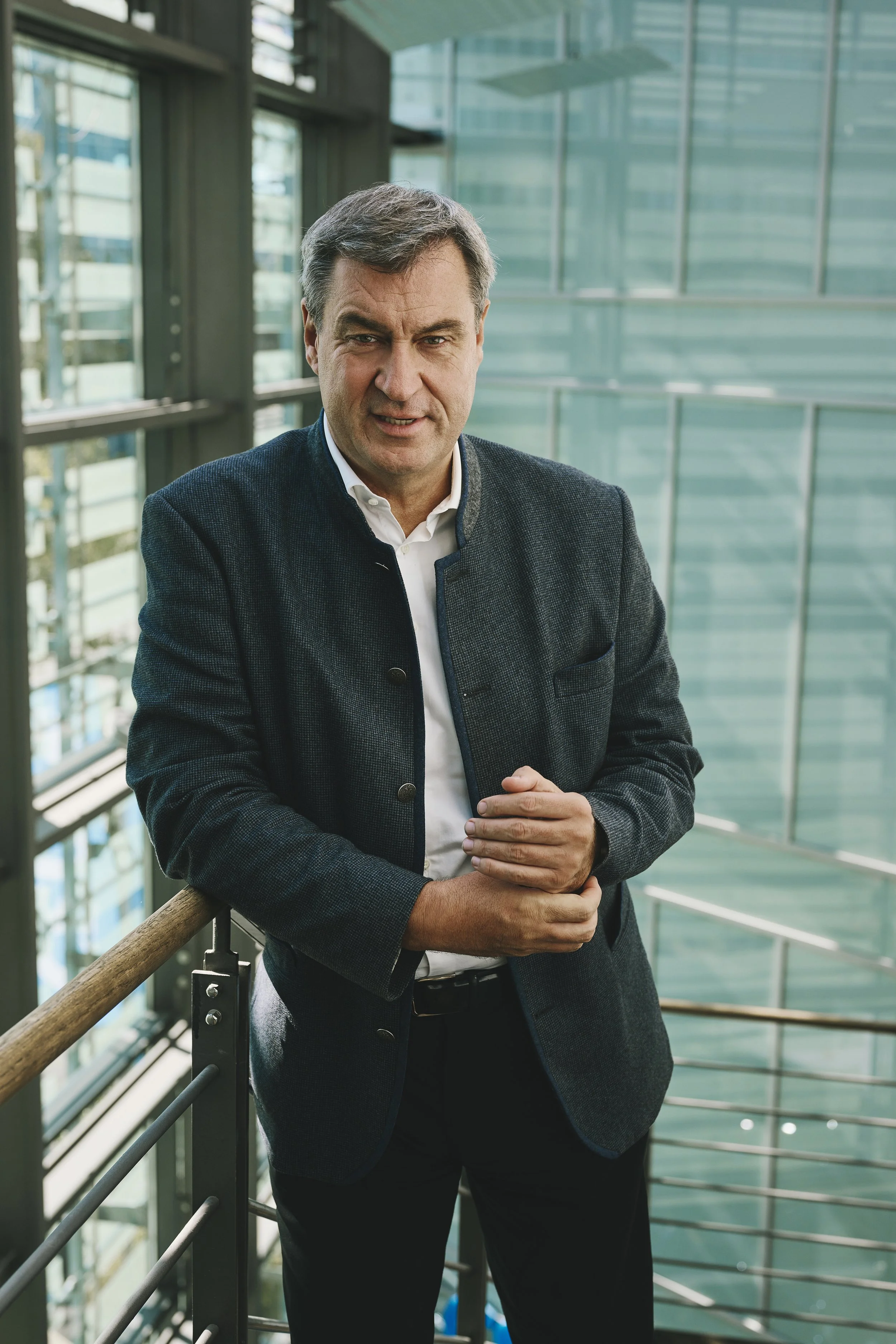 Markus Söder steht in einem modernen Gebäude mit Glaswänden und Metallgeländern. Er schaut zur Kamera und hat die Hände gefaltet.