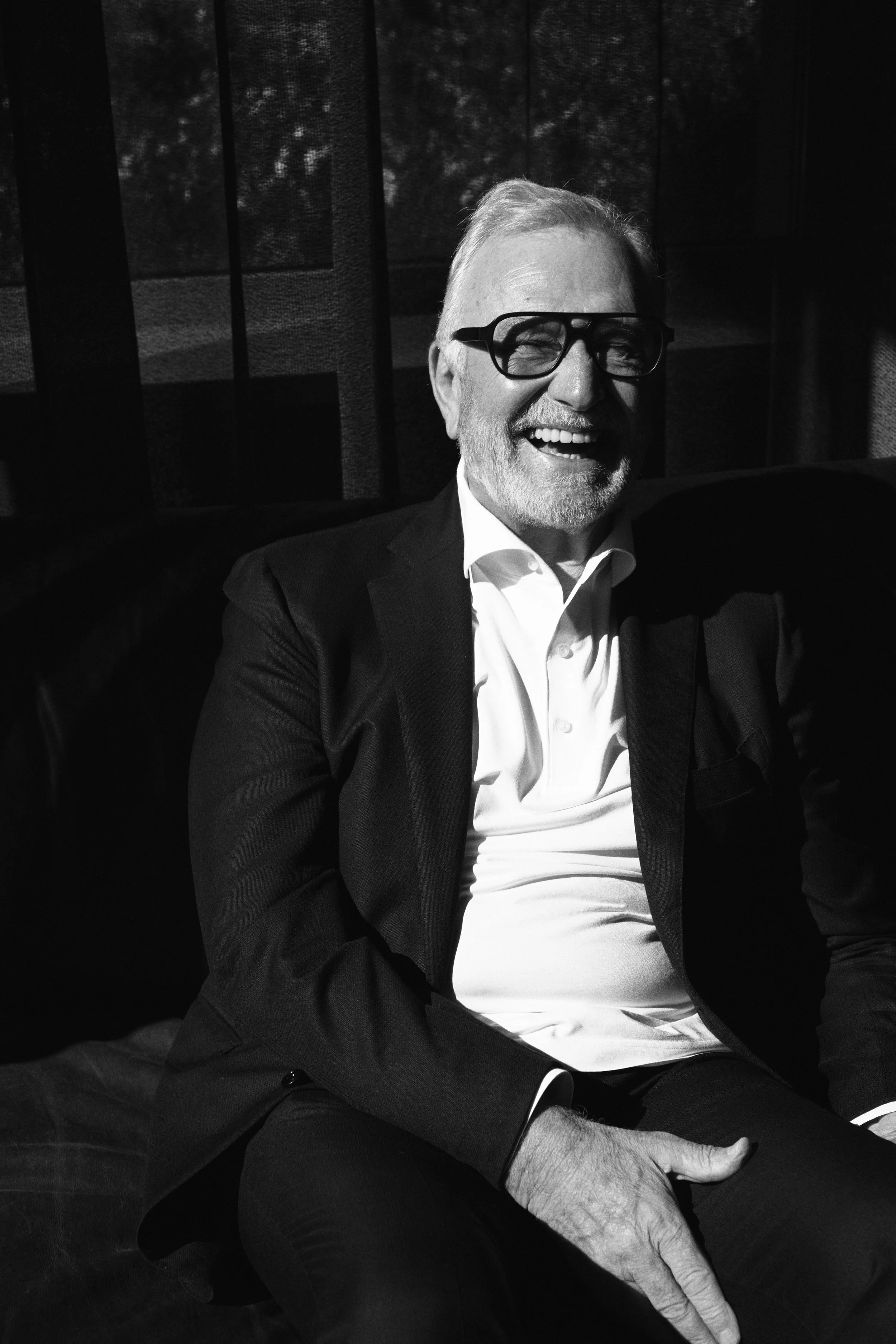 Dieter Müller lacht in einem dunklen Anzug und Hemd. Die Szene ist in Schwarz-Weiß aufgenommen, er sitzt und wirkt entspannt.