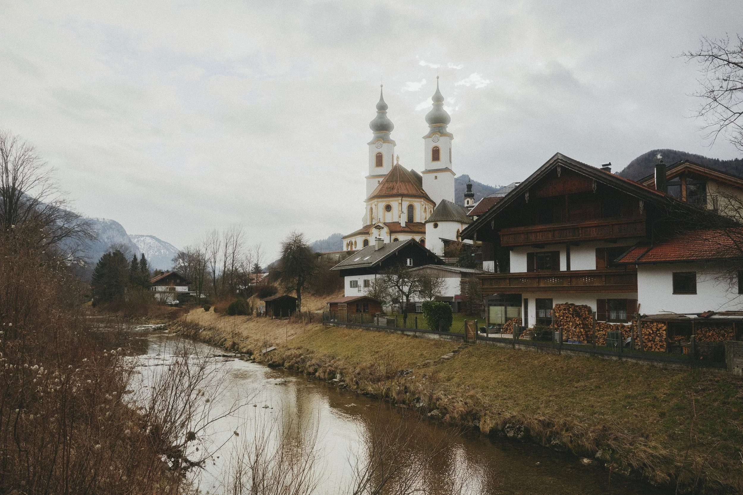 Landschaft in einem bayerischen Dorf mit einer Kirche mit Zwiebeltürmen am Flussufer, umgeben von traditionellen Häusern und Bäumen.