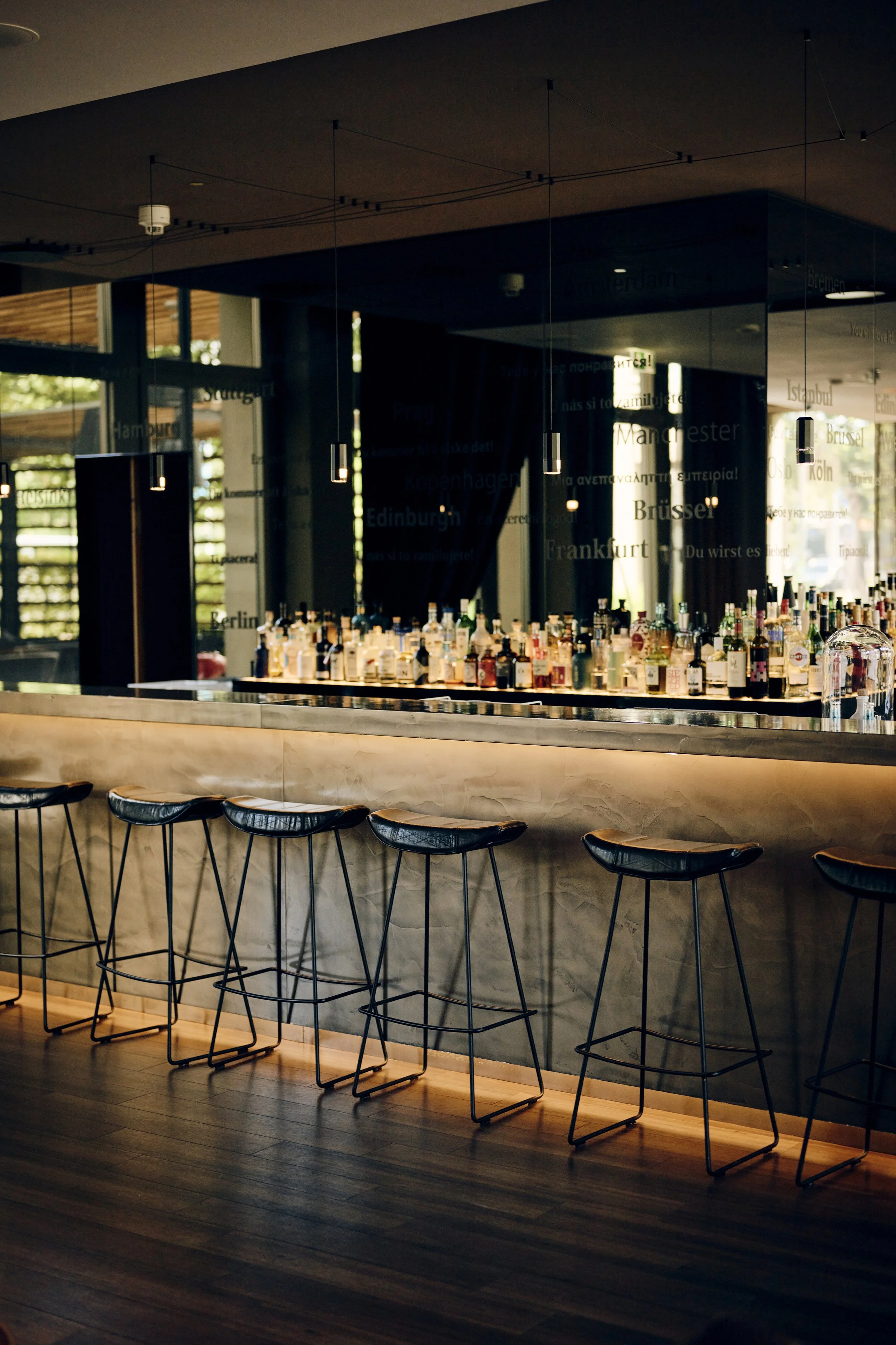 Leere Bar mit mehreren hohen Barhockern vor einer langen Theke, auf der eine Vielzahl von Spirituosenflaschen steht. Hängende Lampen beleuchten den Raum, der modern gestaltet ist.