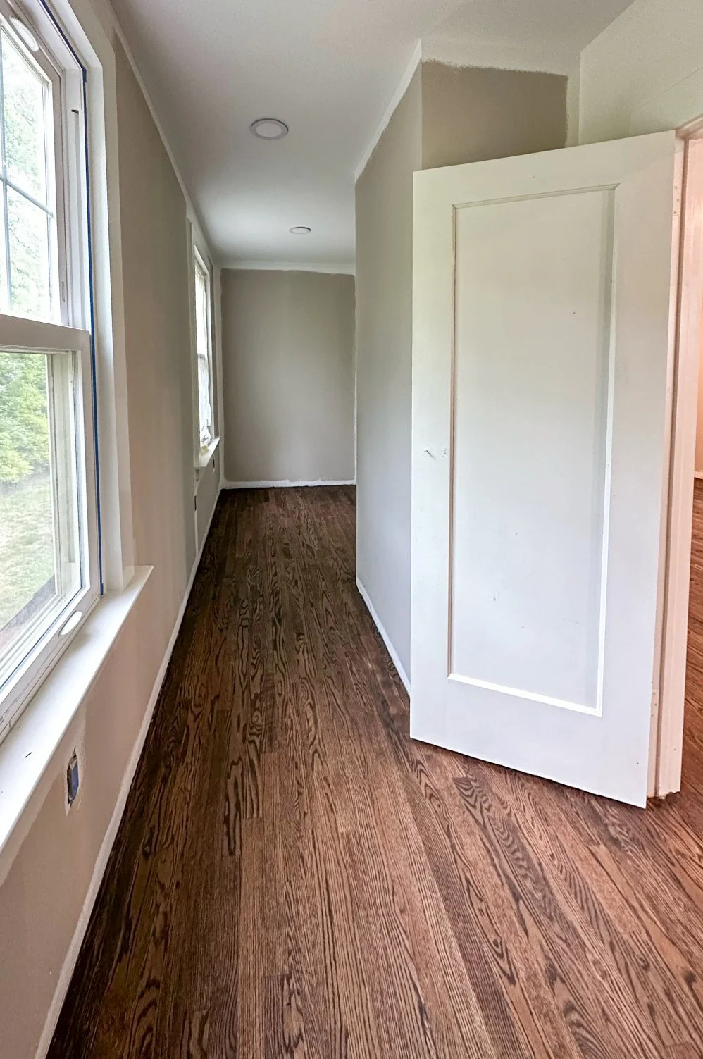 A narrow hallway with wood flooring, beige walls, and multiple windows on the left side. A partially open white door is on the right side.