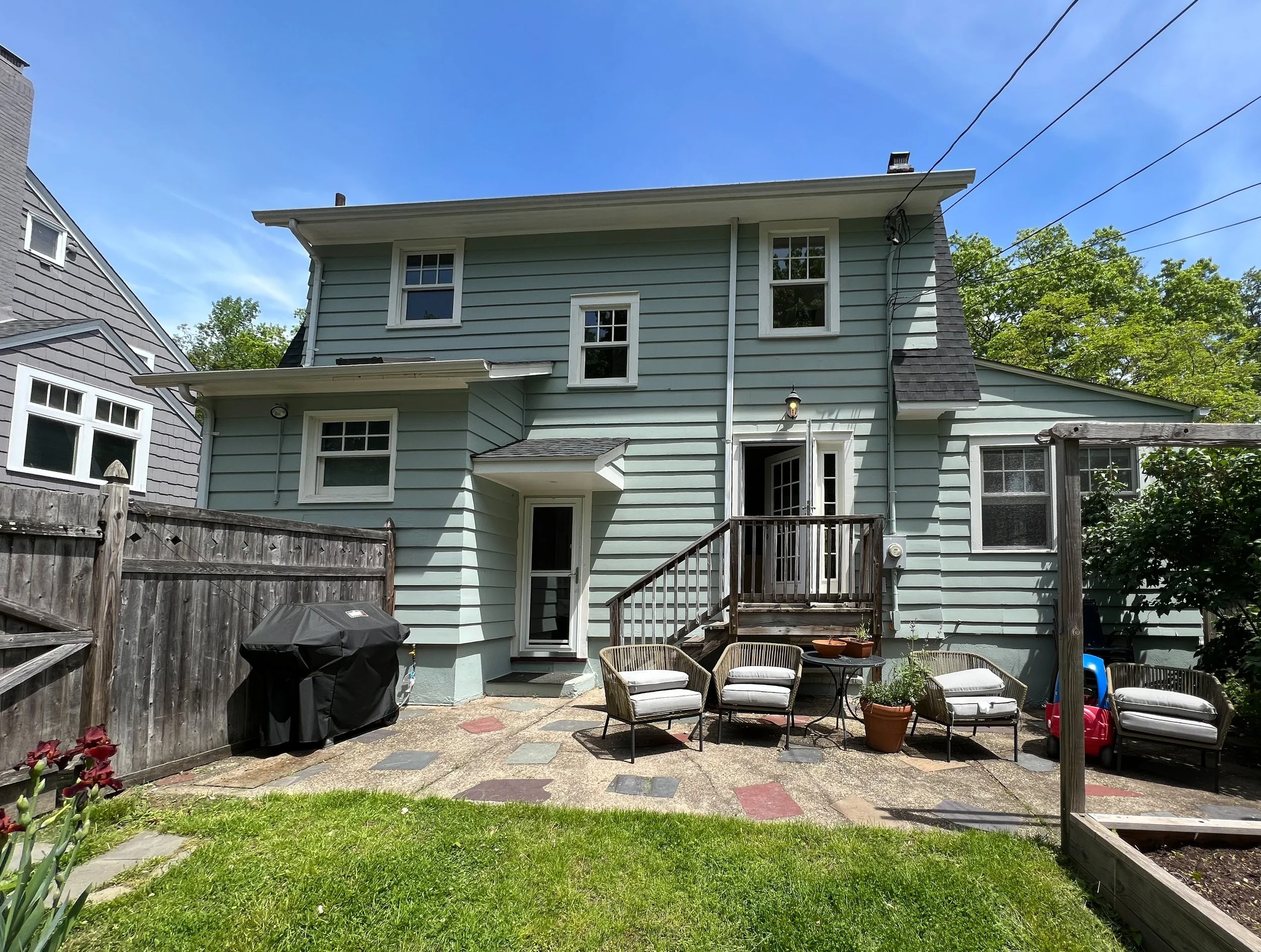 Backyard patio with outdoor chairs, a table, potted plants, a grill, and a two-story light blue house with white trim and a small staircase.
