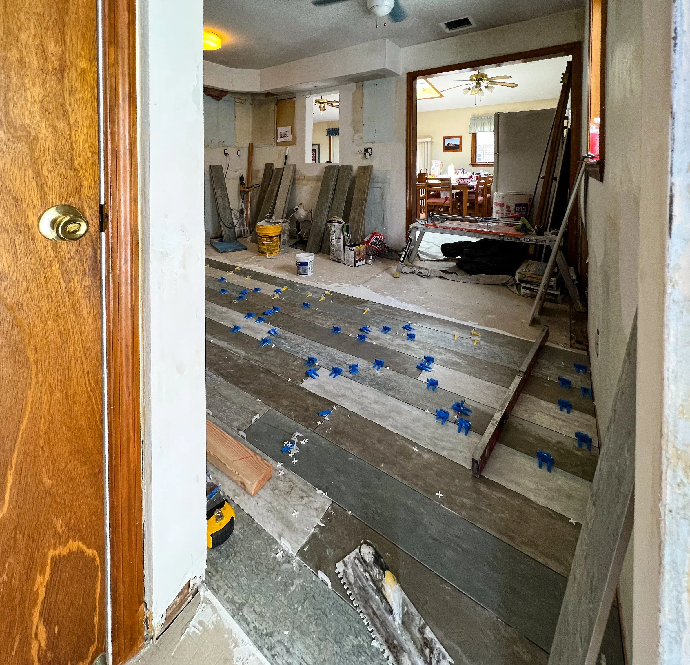 Interior of a house under renovation with subflooring being installed, blue and yellow tile spacers scattered on the floor, construction tools and supplies, and rooms visible in the background.