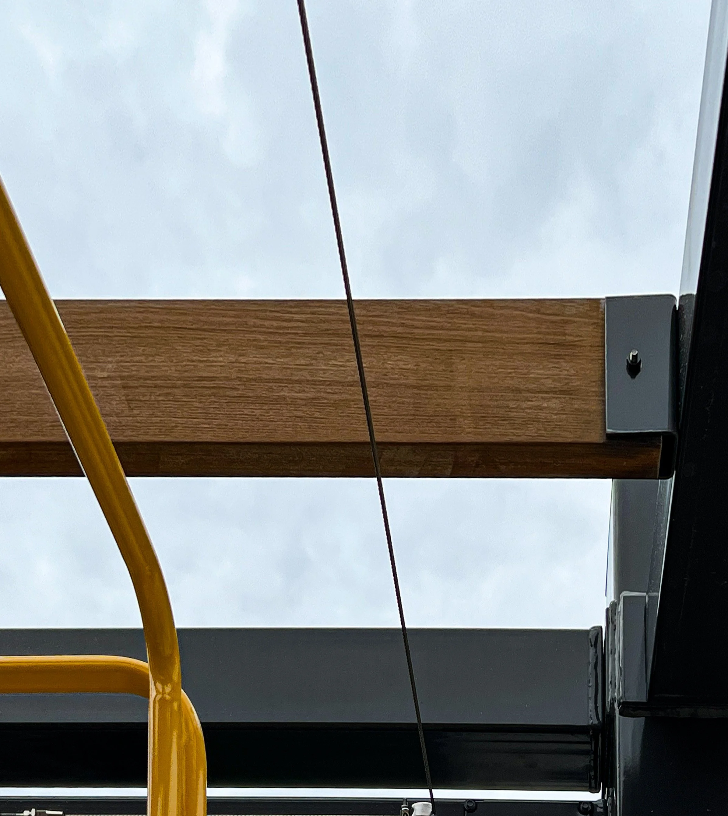 Close-up of a corner of a wooden table or bench with metal brackets, a yellow pole, and a horizontal black cable against a cloudy sky.