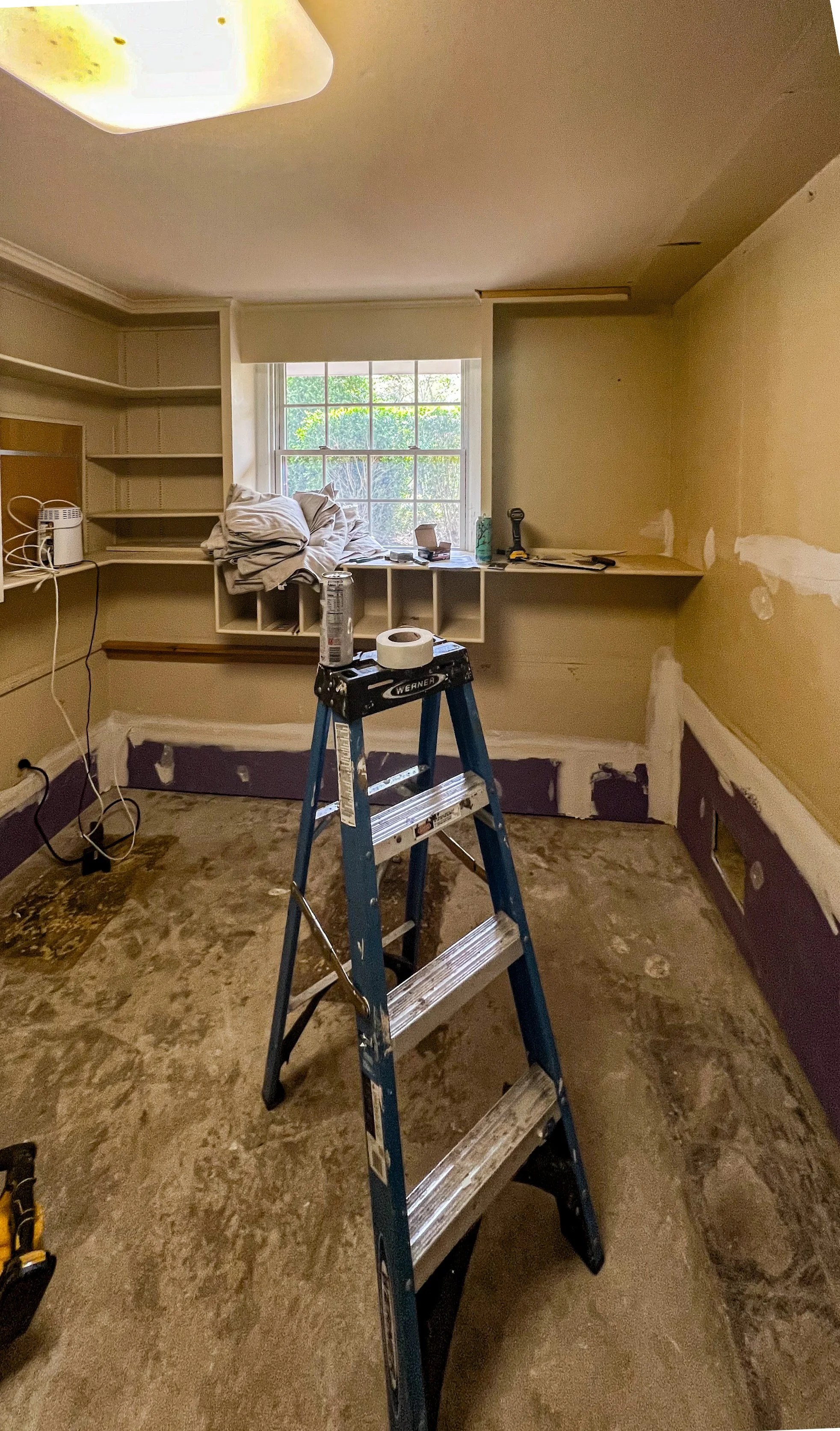 A room under renovation with unfinished walls, a ladder with paint cans and supplies, and a window with a view of greenery outside.