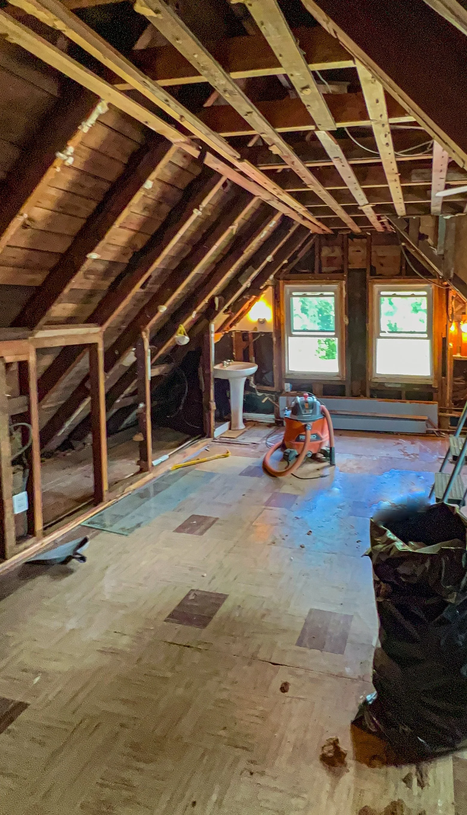 A partially renovated attic with exposed wooden beams and unfinished walls, a small sink, a vacuum cleaner, and two windows