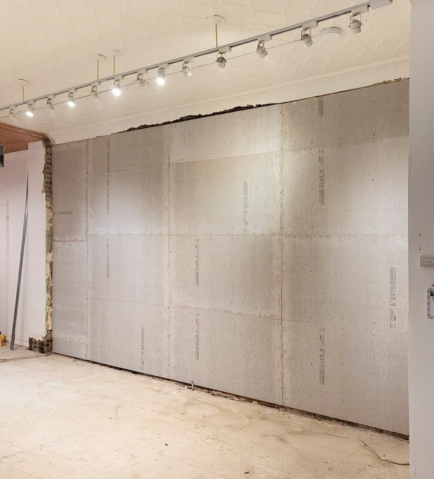 Interior wall under construction with drywall panels installed, ceiling lighting fixtures overhead, and unfinished flooring.