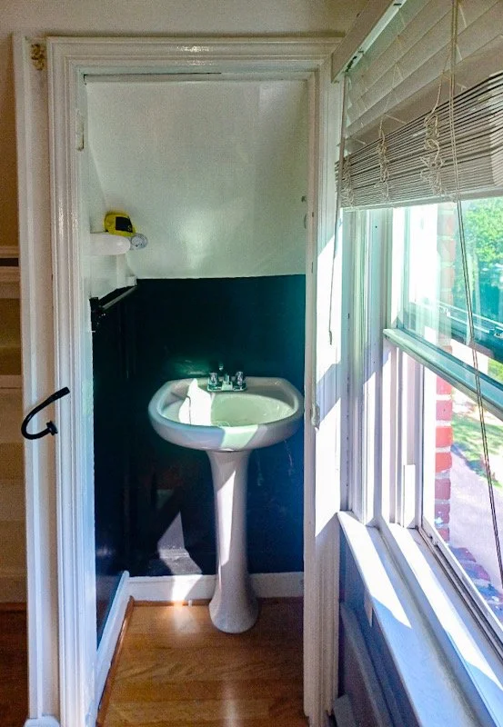 Small bathroom with a white pedestal sink, dark blue wainscoting, and white walls. A window with partially open blinds is on the right, letting in natural light.