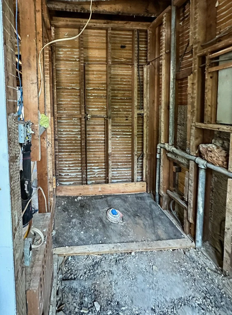 Interior of a bathroom under renovation, showing exposed wooden framing, plumbing pipes, and a rough concrete floor with debris.