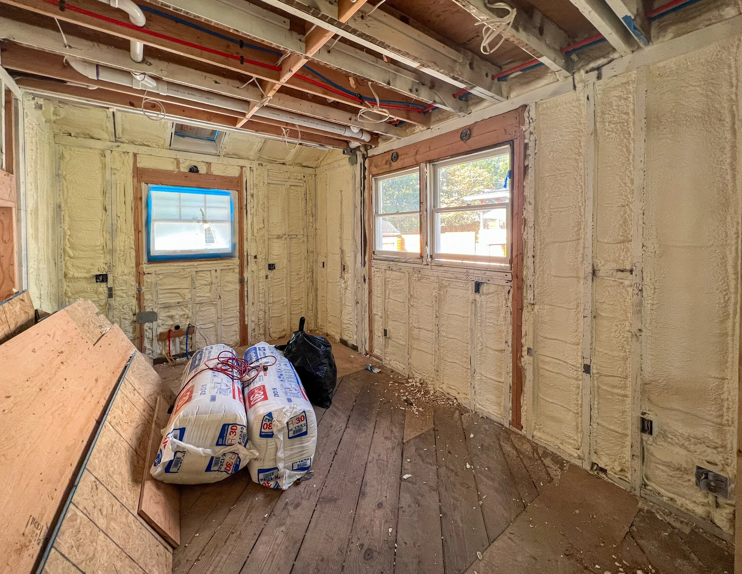 Room under renovation with exposed insulation, wooden floor, and construction materials, including insulation bags and plywood sheets.